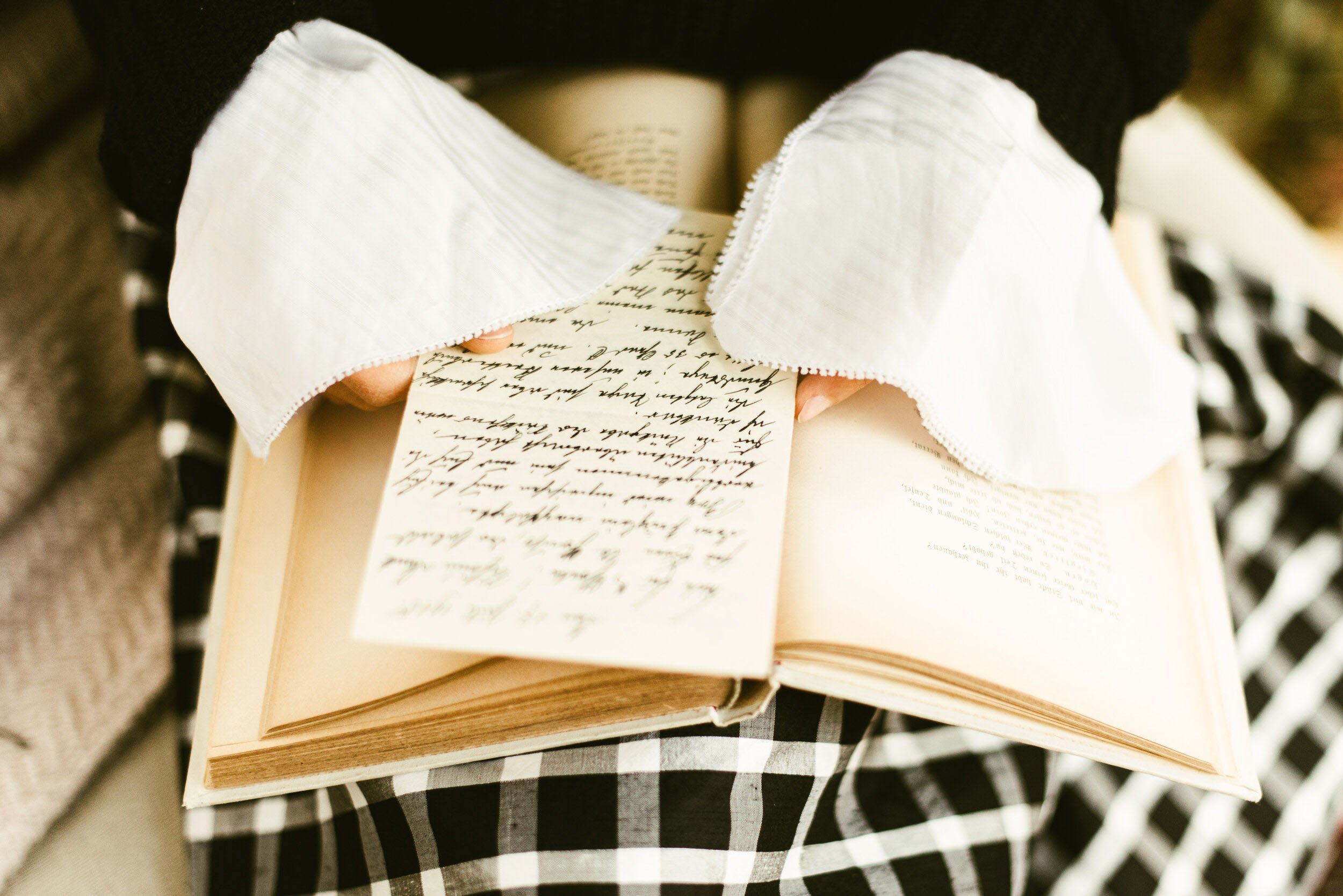 A person holding an open book with handwritten text, sitting on a checkered fabric surface, with a white paper or note partially visible underneath.