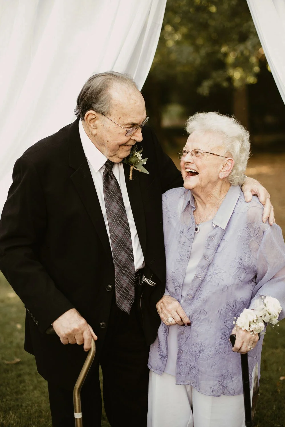 An elderly man and woman sharing a joyful moment outdoors during a special occasion, with the man in a black suit and the woman in a light purple blouse, both using walkers.