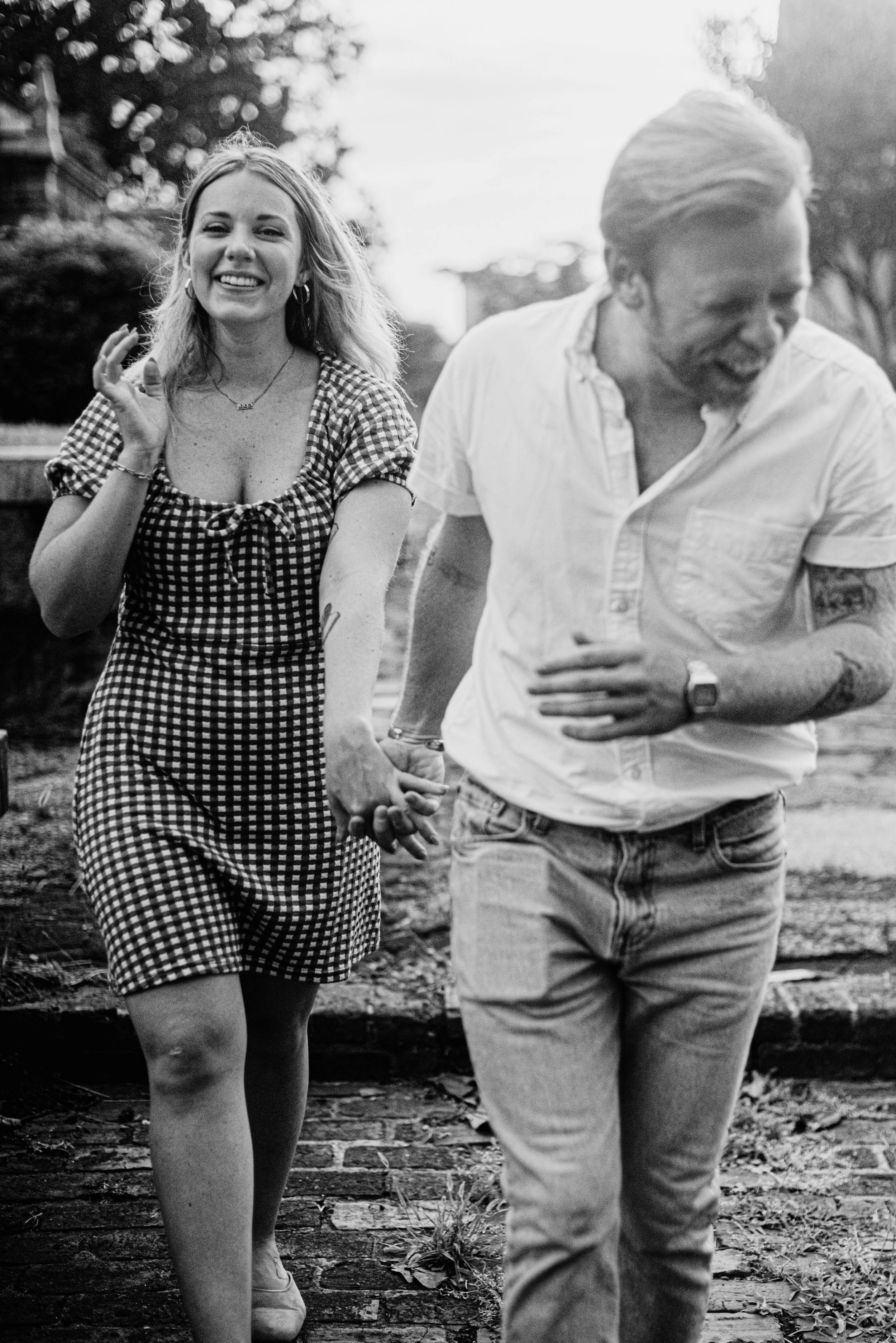 A black and white photo of a smiling woman holding hands with a man who is looking down and laughing, walking outdoors on a wooden pathway.