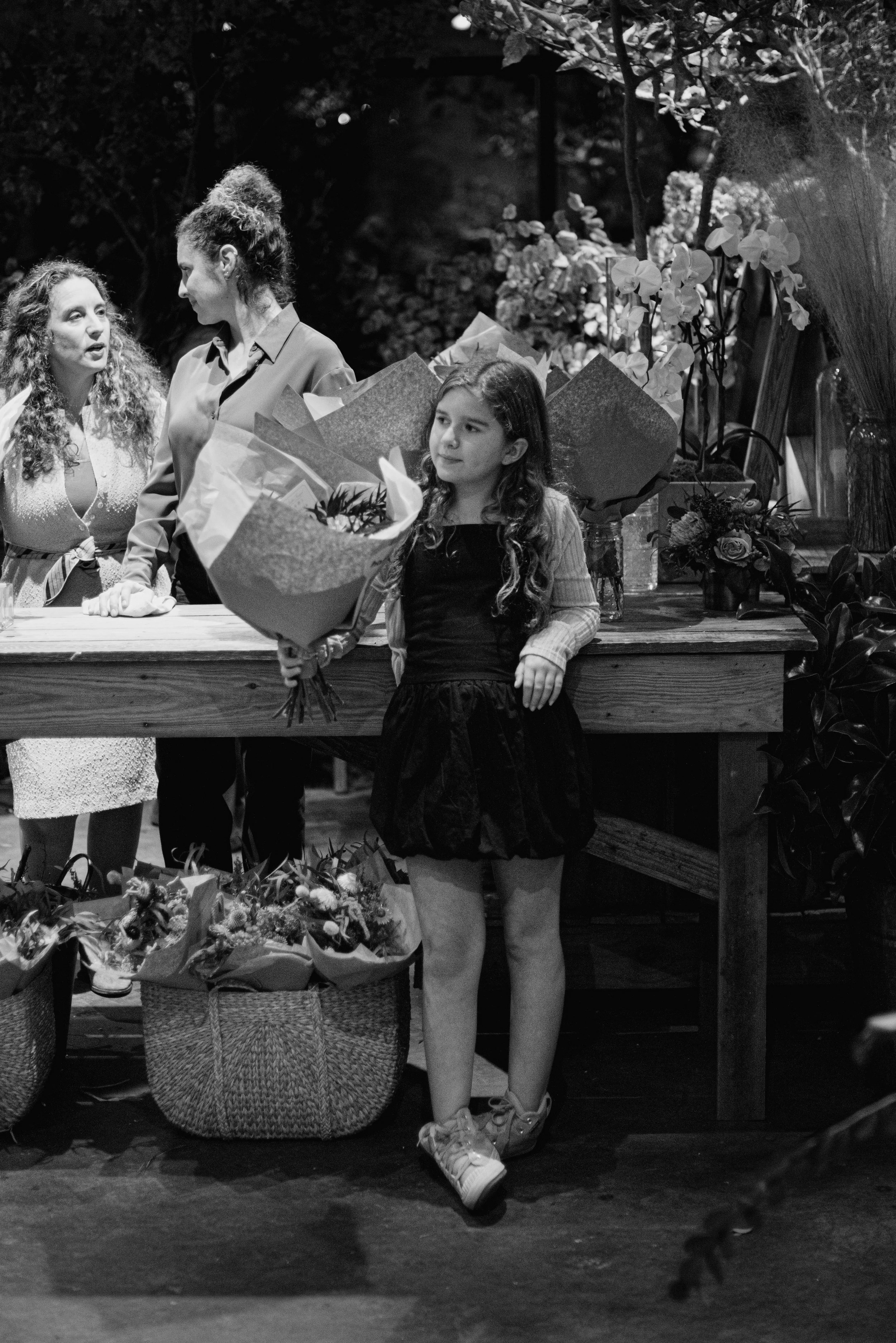 Three women at a flower shop. One woman with long, curly hair and a light dress is talking to another woman with curly hair and a bun. A young girl with long hair wearing a jacket and skirt is holding a large bouquet of flowers. There are baskets of 