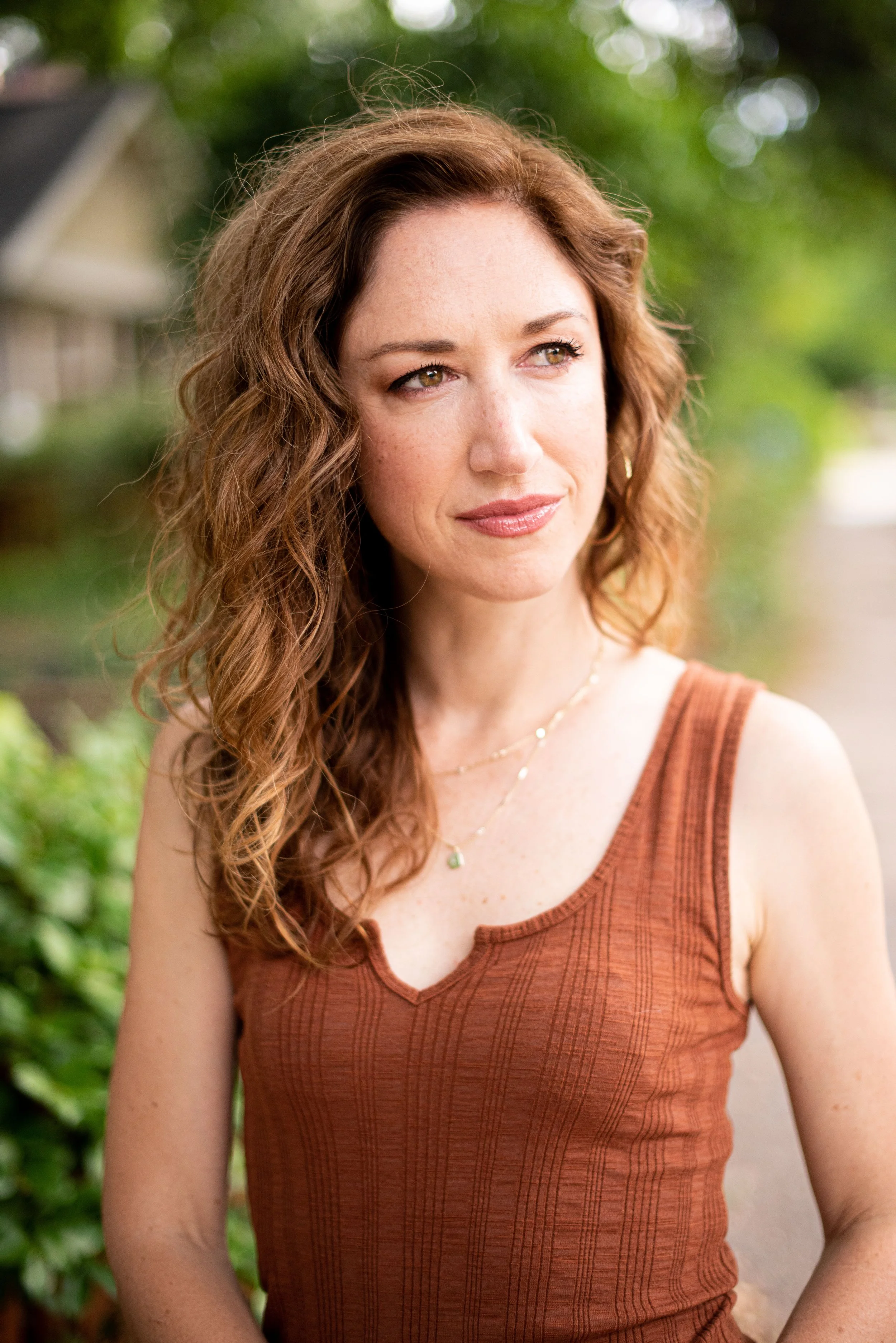 A woman with curly auburn hair and light skin is standing outdoors, wearing a rust-colored tank top and layered necklaces. She is looking off to her left with a gentle smile, with a blurred background of greenery and a house