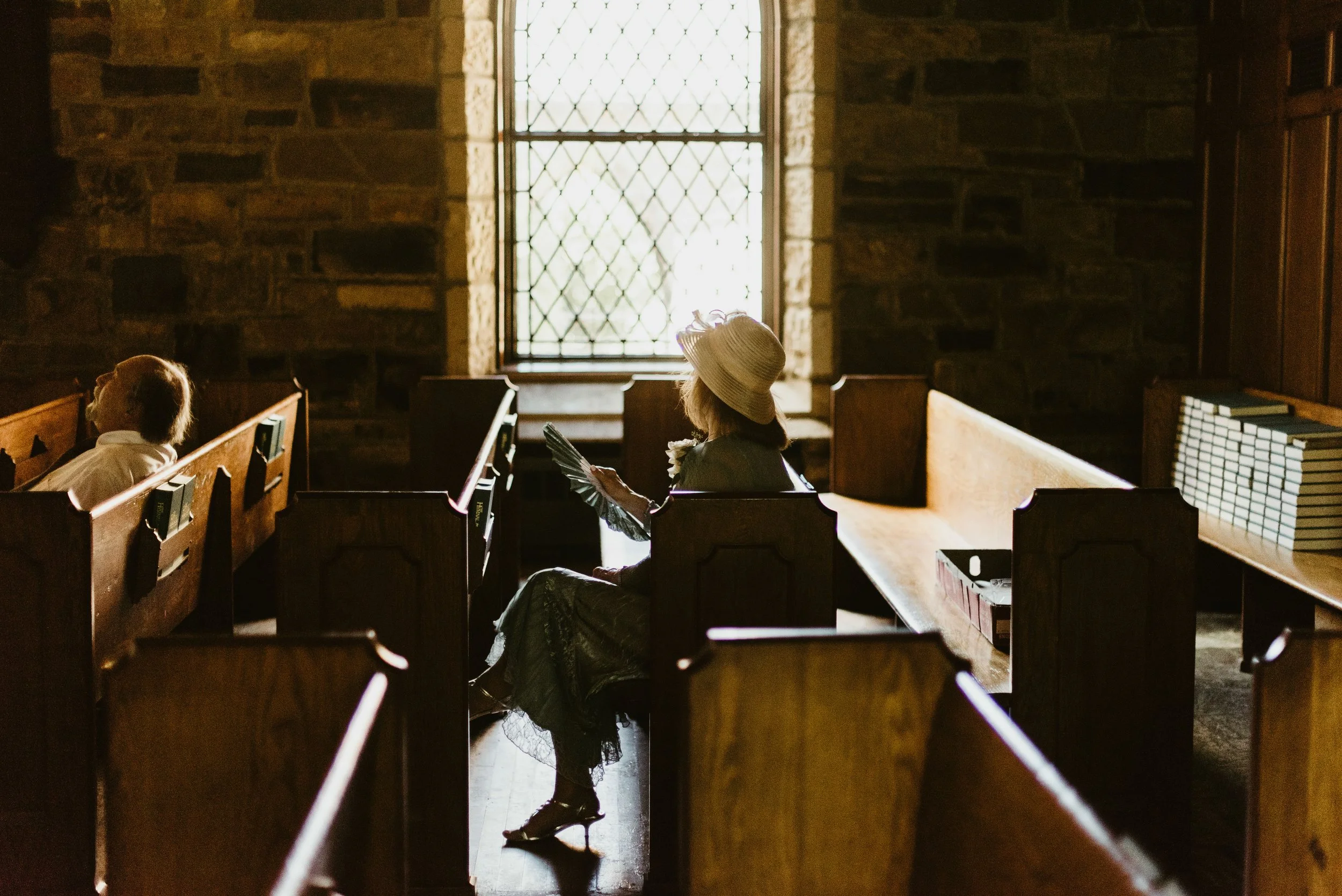 Two women sitting in a church pew, one wearing a wide-brimmed hat and black dress holding a fan, the other with short hair looking upward, with a stained glass window and stone wall behind them.