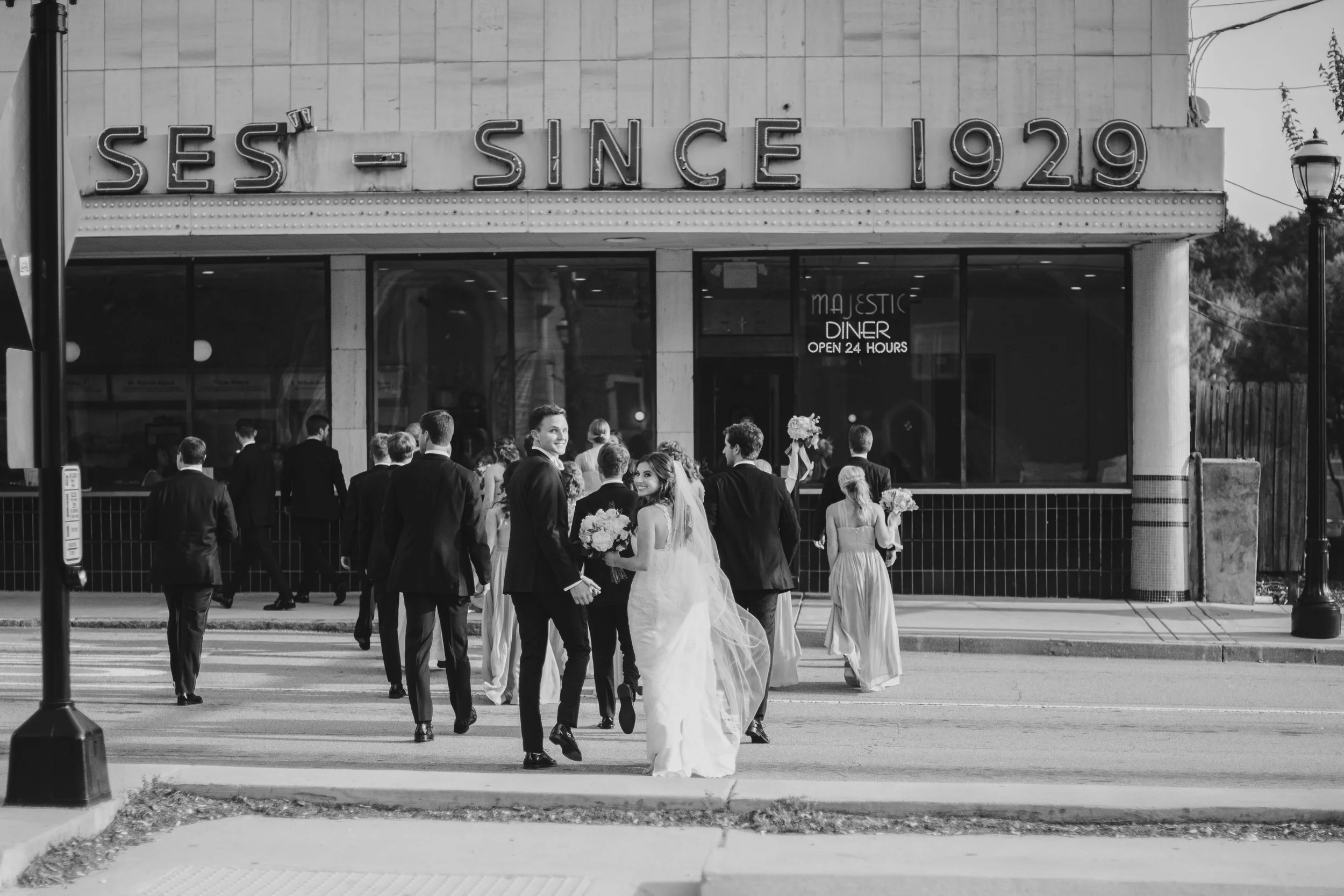 A wedding procession with a bride holding a bouquet and surrounded by guests, walking away from the camera outside a building with a sign that reads 'Ses - Since 1929' and 'Majestic Diner, Open 24 Hours'.