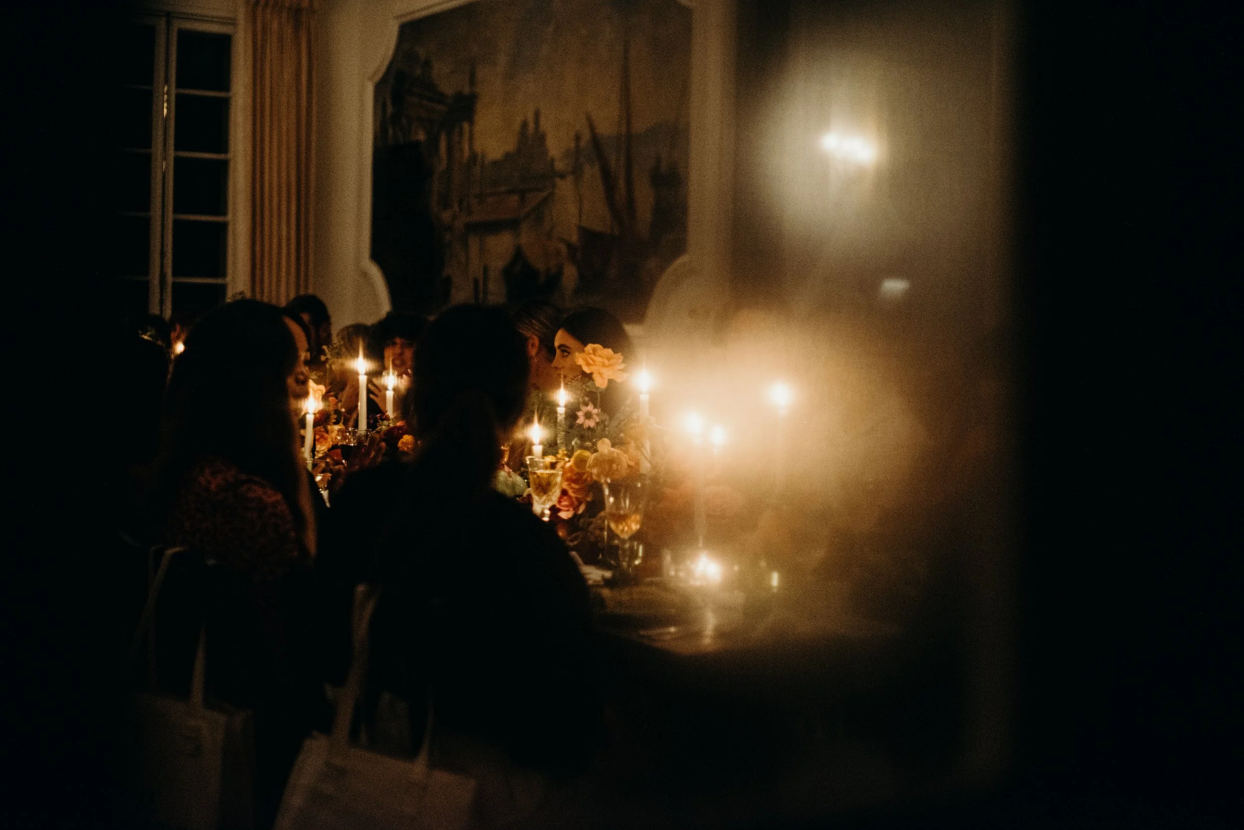 A dimly lit indoor party with people sitting at a long table decorated with flowers and candles, with a large framed painting on the wall in the background.