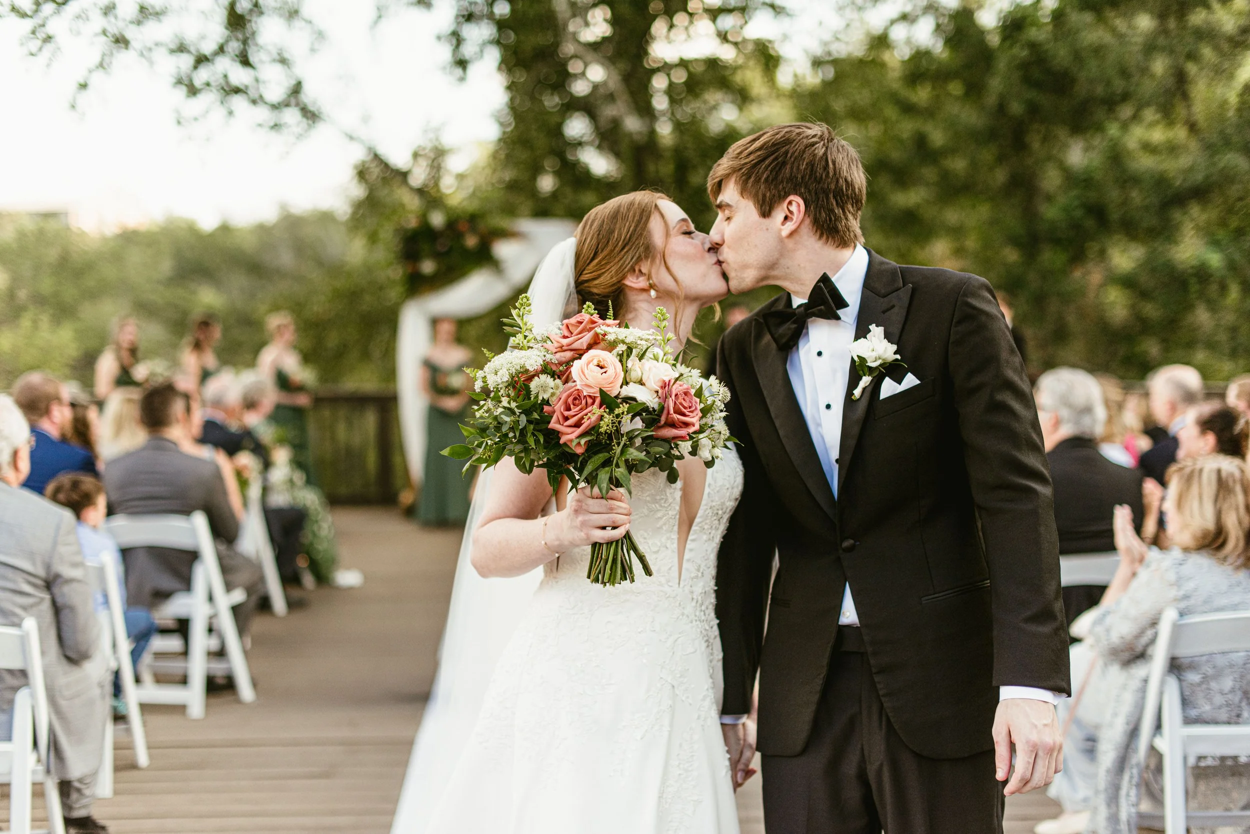 A bride and groom kiss during an outdoor wedding ceremony, with guests seated in the background.