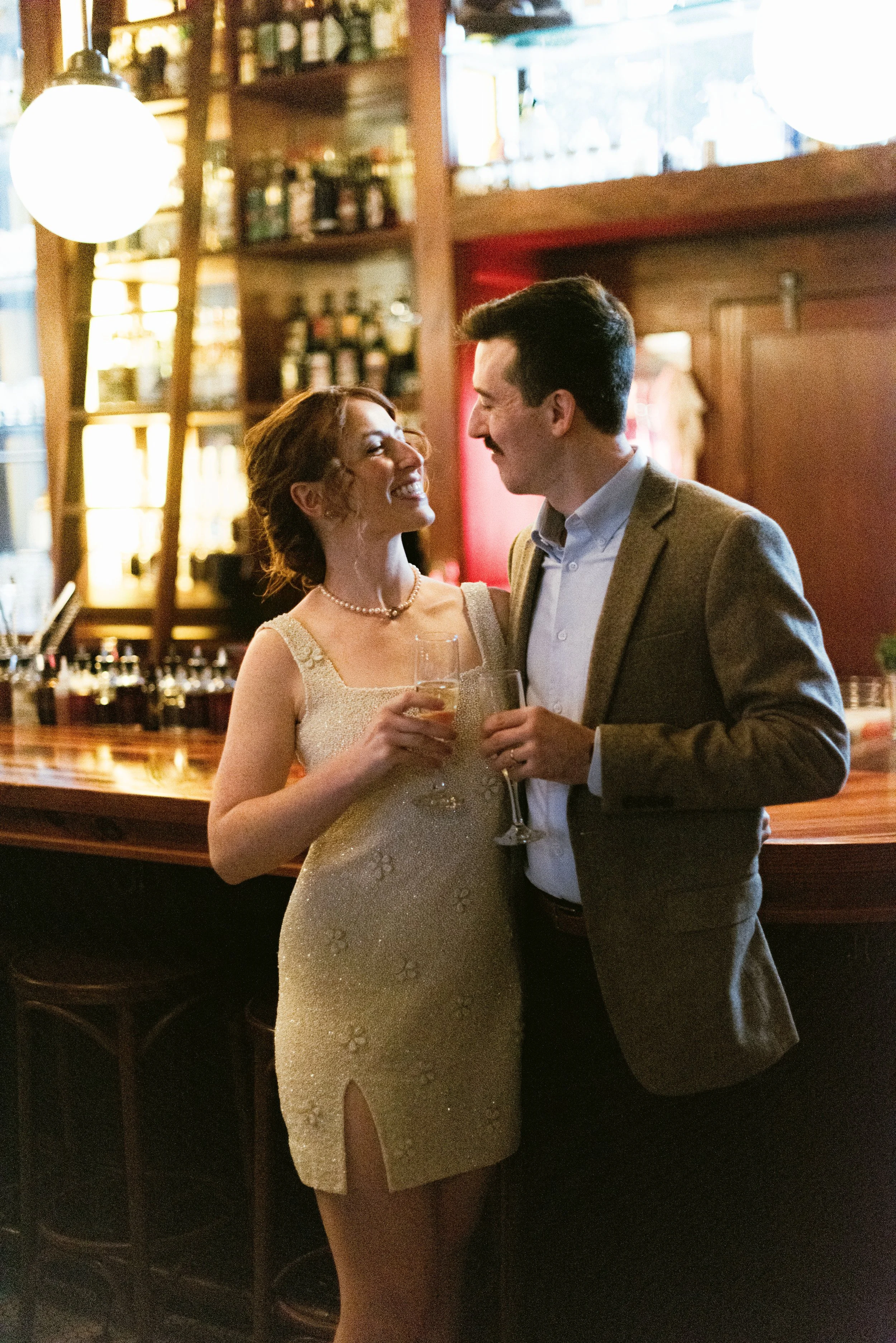 A woman in a white, sparkly dress and a pearl necklace, holding a glass of champagne, smiling at a man in a gray blazer and white shirt. They are in a warmly lit bar or restaurant setting with wine bottles on shelves in the background.