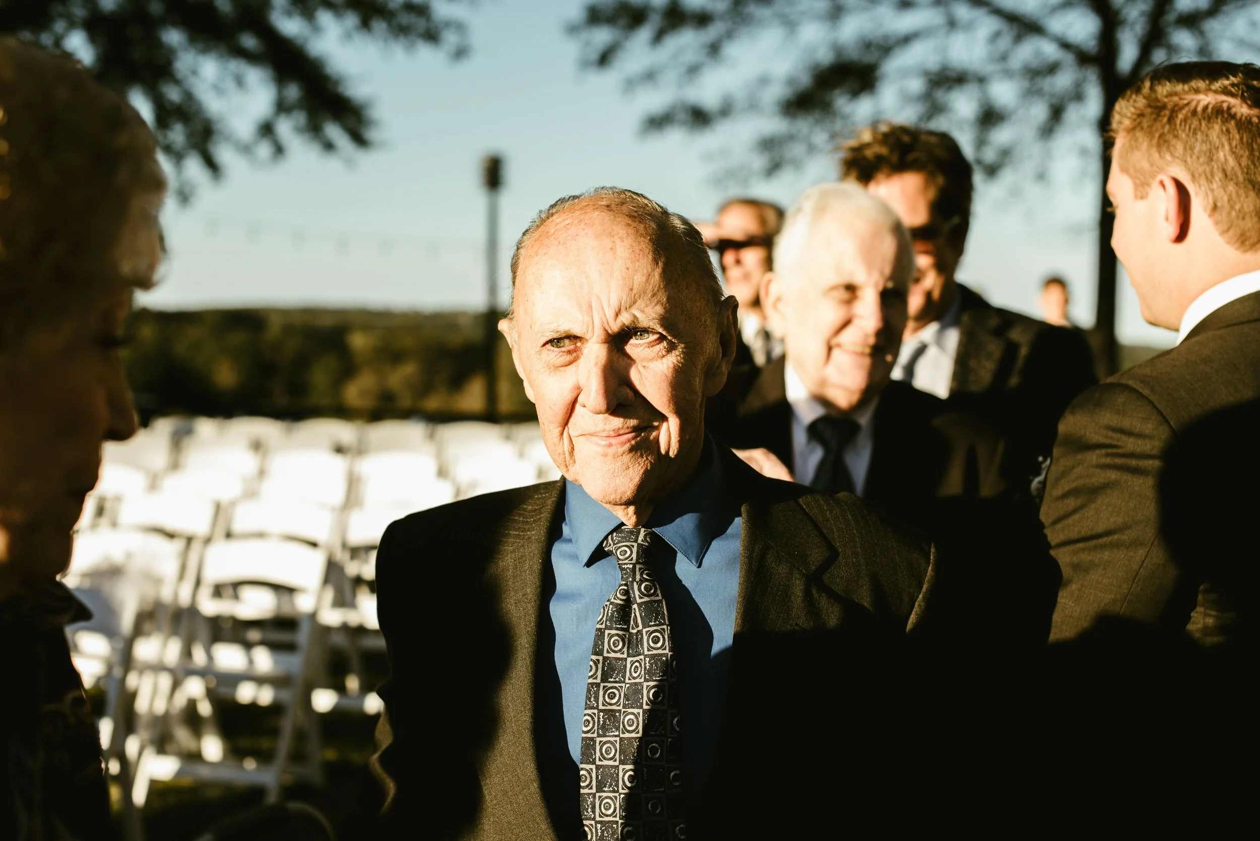 A group of people dressed in formal attire, sitting outdoors during daytime, with a focus on an older man in a black suit and patterned tie, engaging in conversation with others around him.