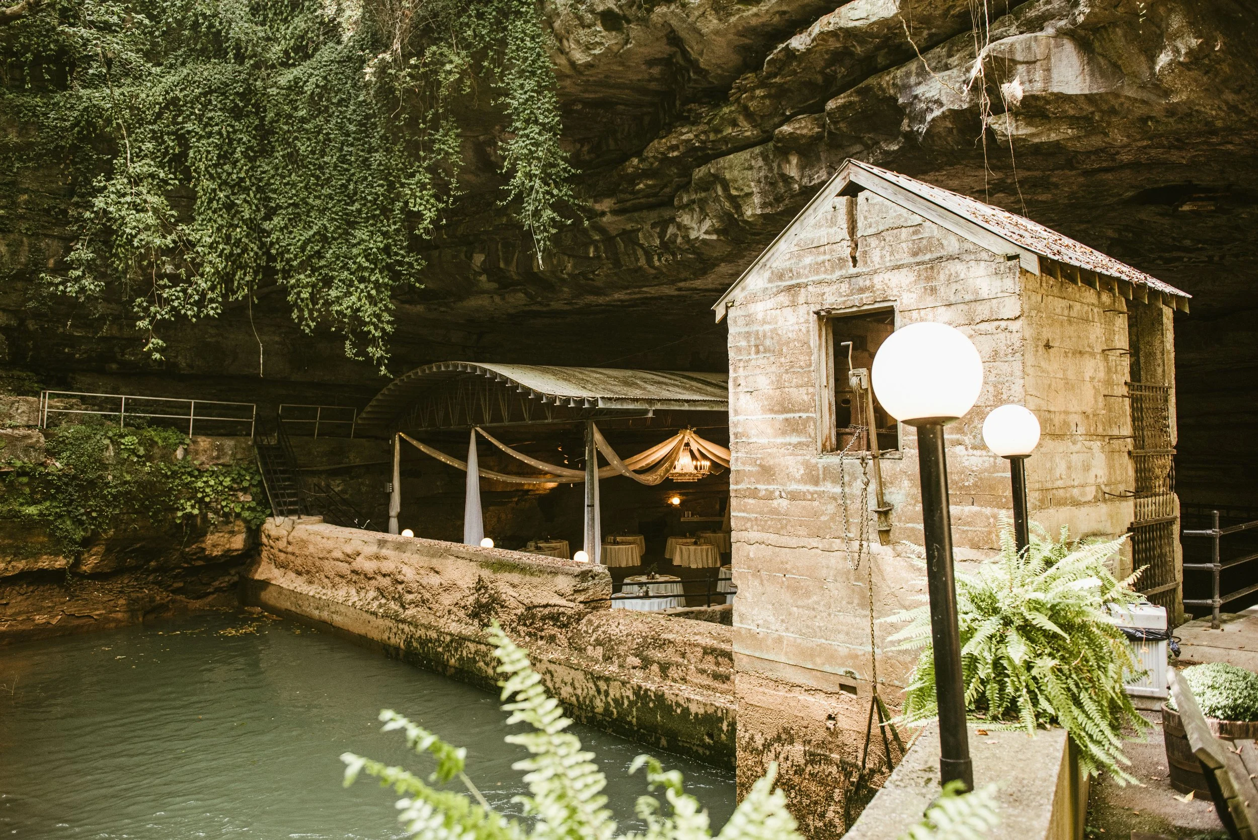 A small concrete building with a sloped metal roof and a window, situated near a water features inside a cave or cavern. There are two spherical lamps in front of the building. Tables with tablecloths and chairs are seen beneath a draped canopy nearb