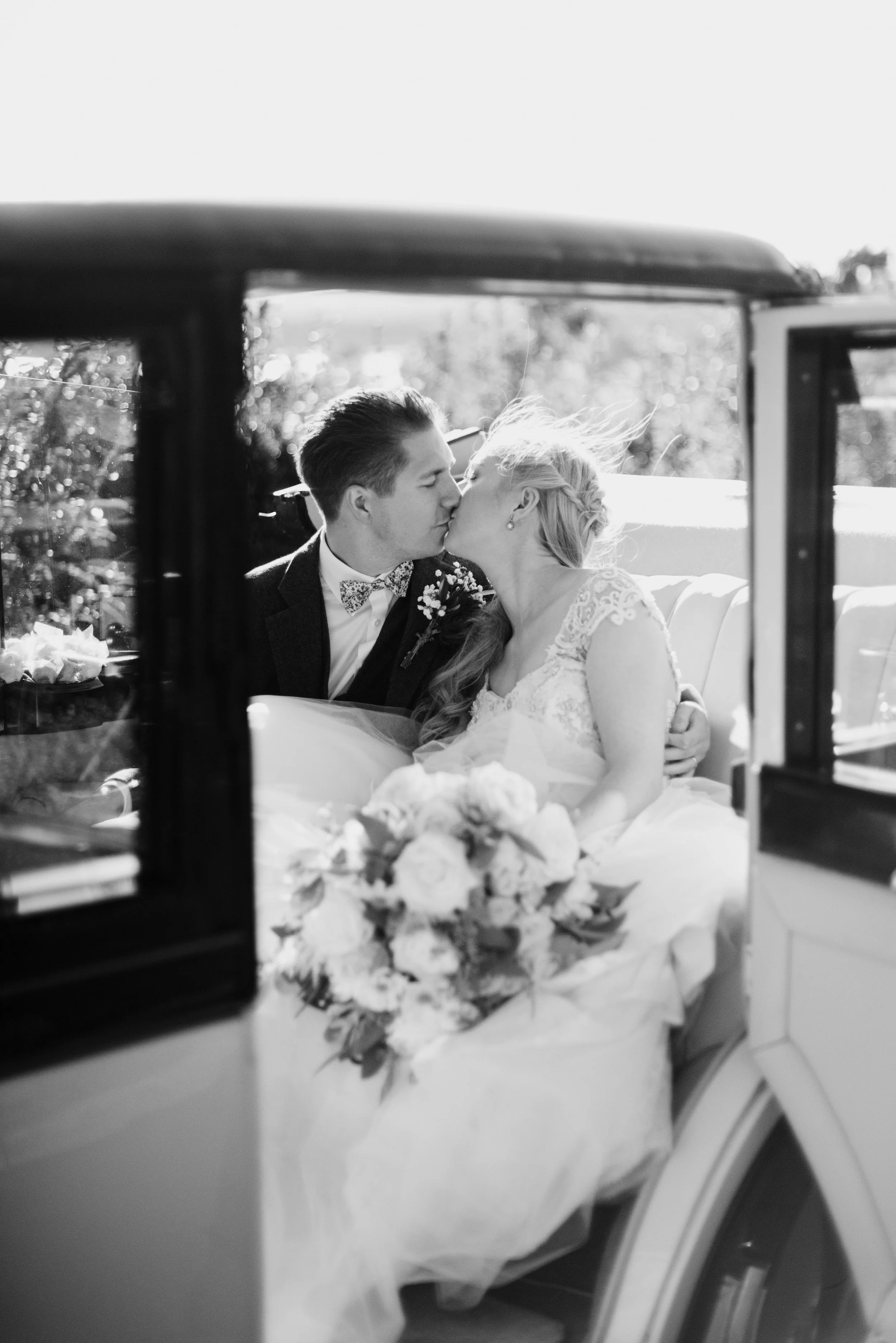 A black and white photo of a bride and groom sharing a kiss inside a vehicle, seen through a window. The bride has braided hair, and the groom is wearing a suit with a bow tie. Flowers are on the table in front of them.