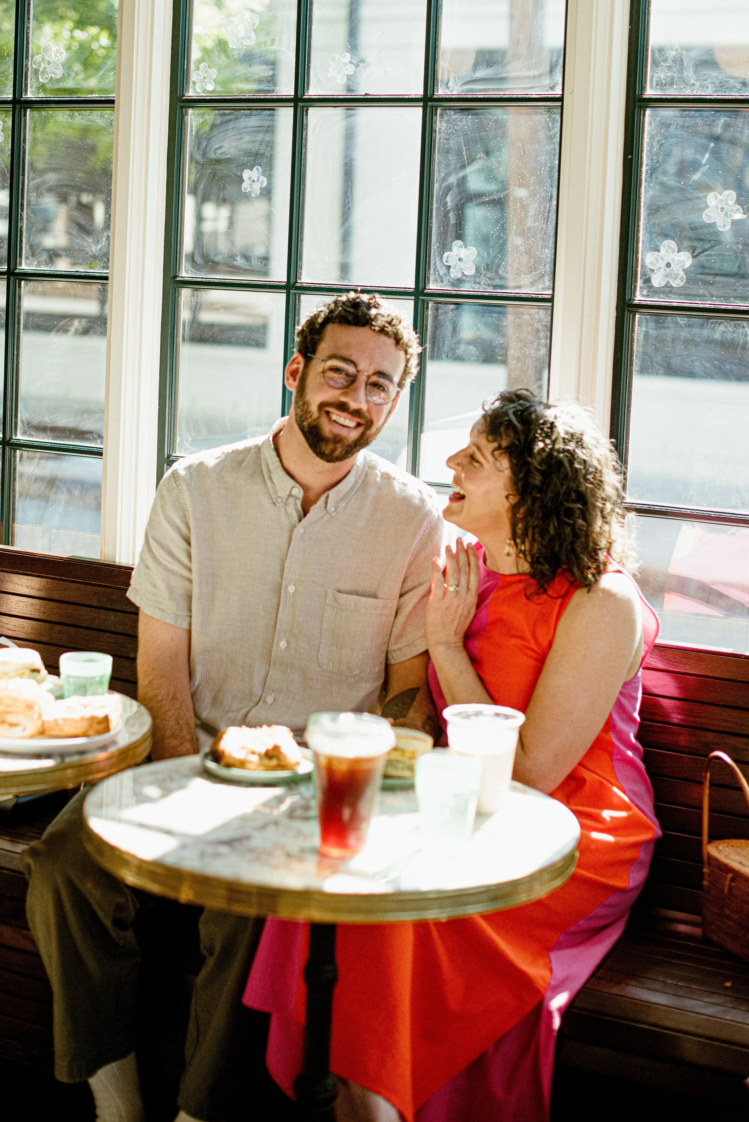 A man with glasses and a beard is sitting at a table, smiling, with a woman with curly hair laughing and touching his shoulder. They are in a sunny room with large windows decorated with flower stickers. The table has various desserts and drinks, and