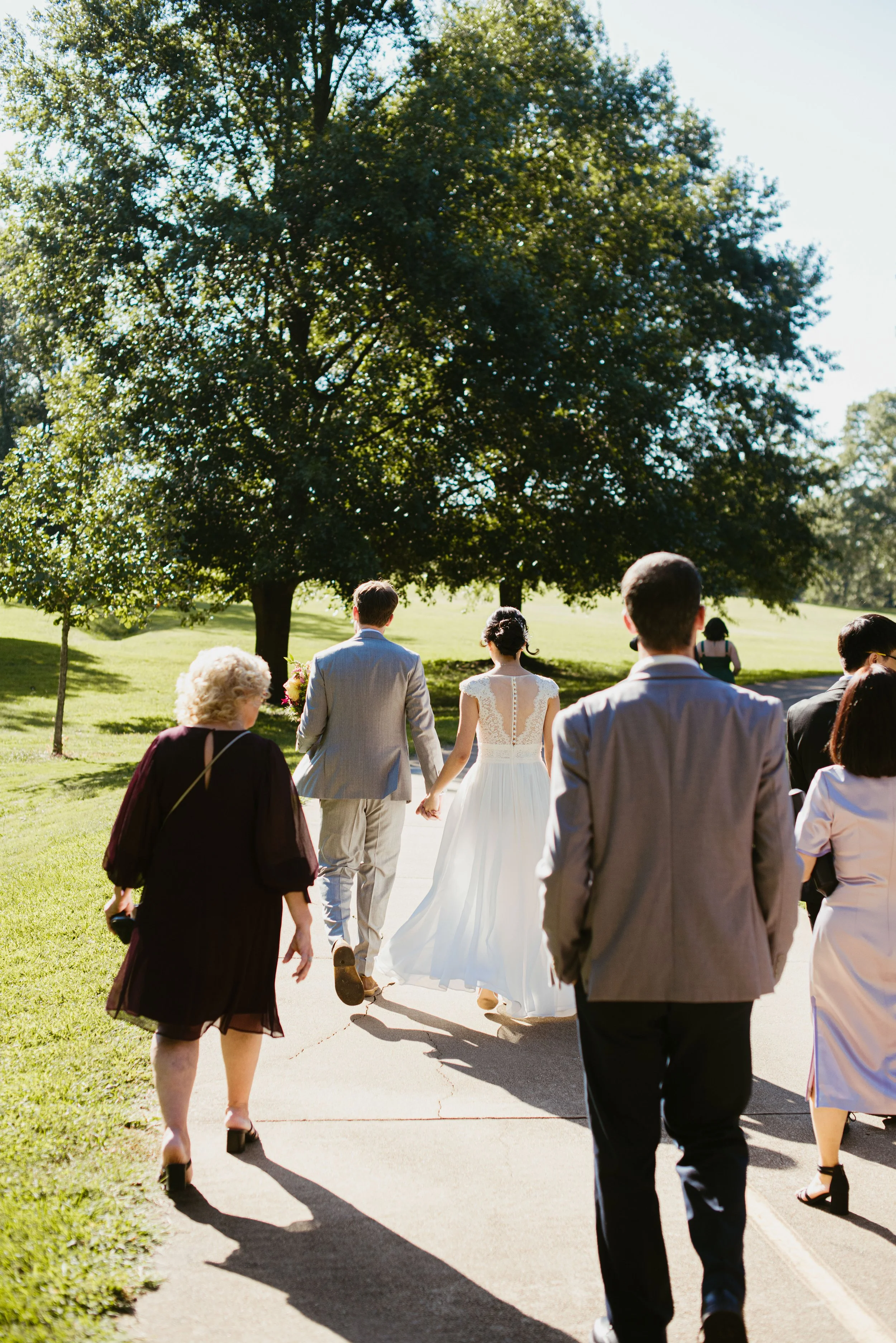 A group of people walking outdoors on a sunny day, with a bride in a white wedding dress leading the way in front of a large green tree.