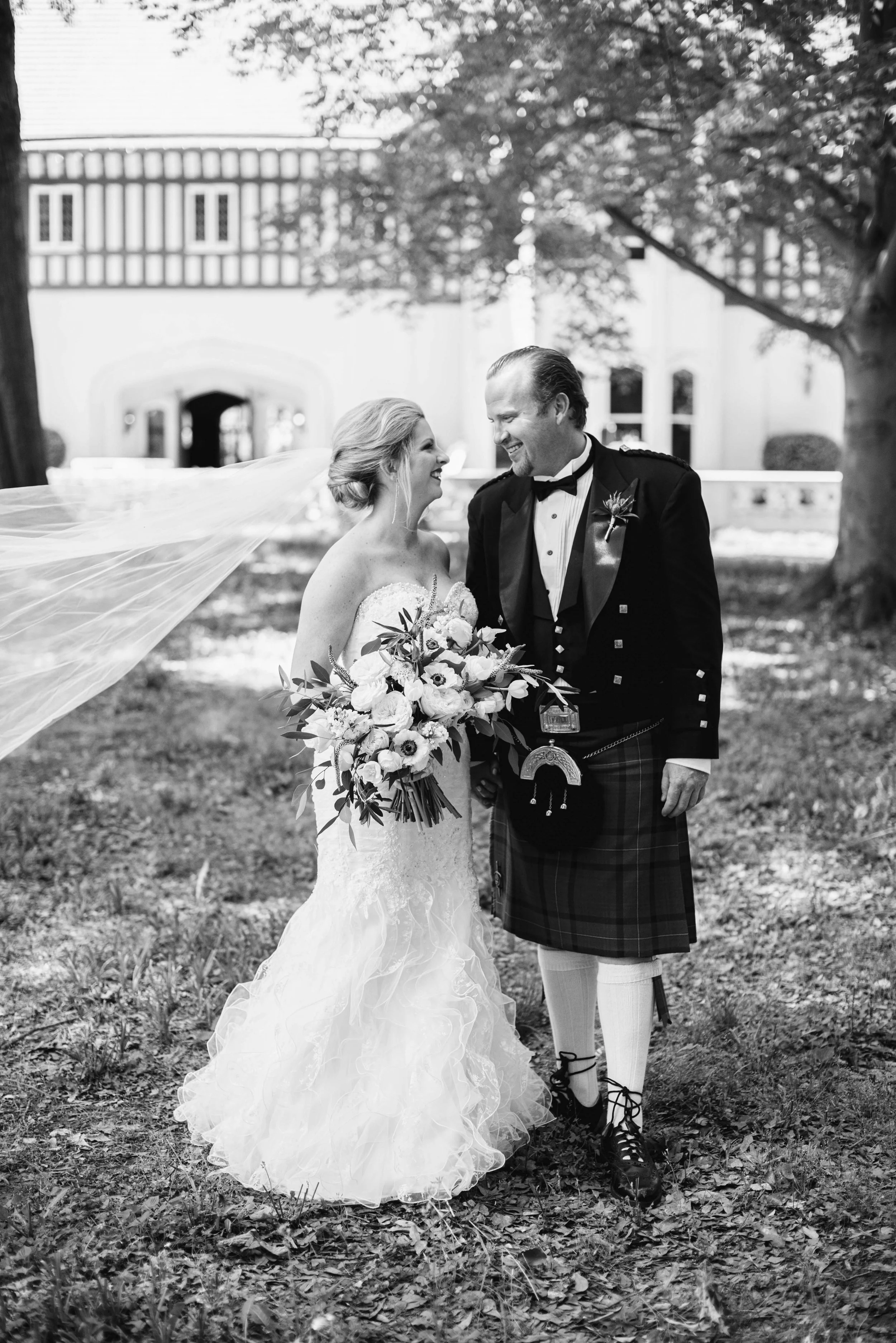 A bride and groom in wedding attire standing outdoors, smiling at each other, with trees and a building in the background.