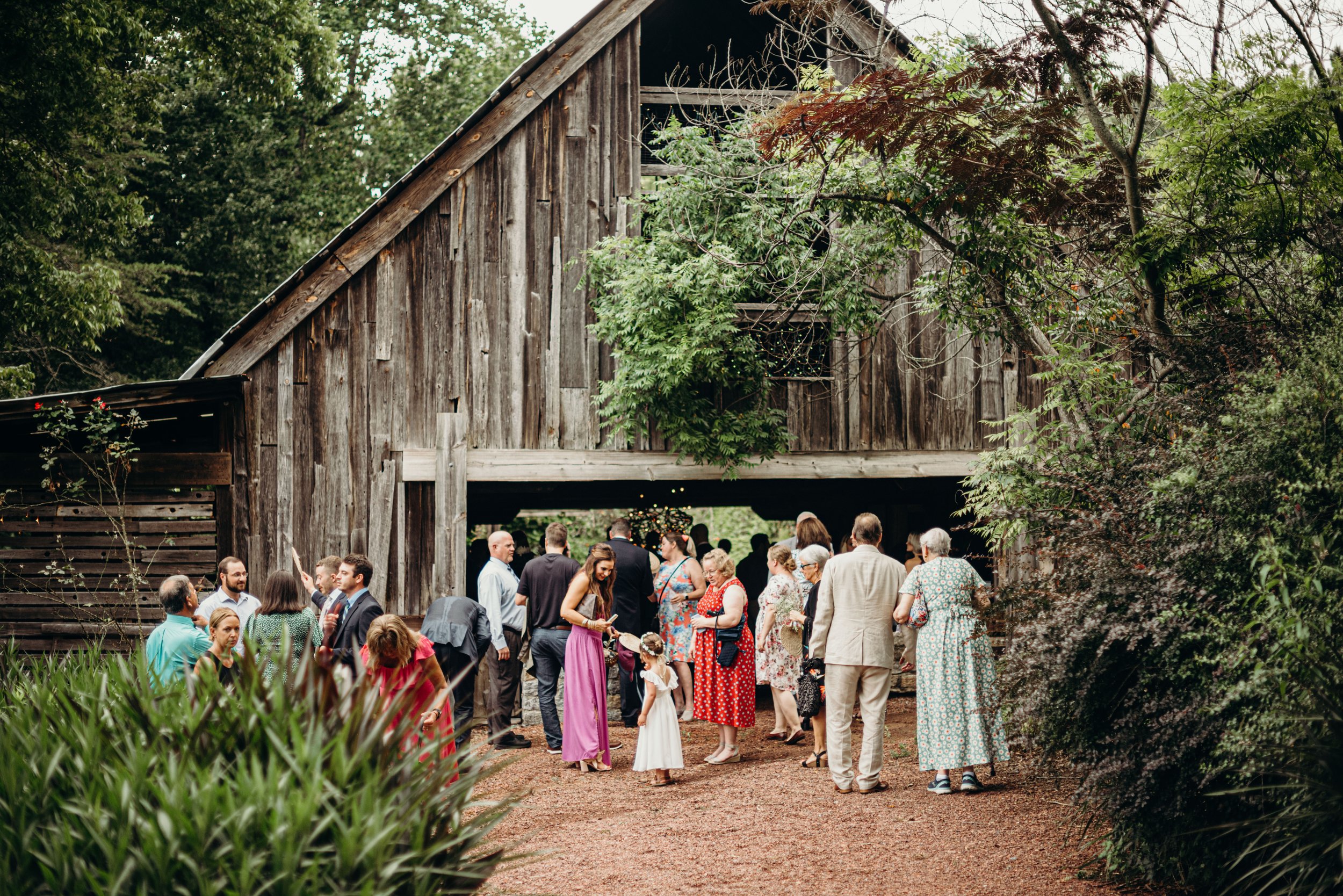 People gathered at an outdoor event by a rustic wooden barn surrounded by trees and greenery.