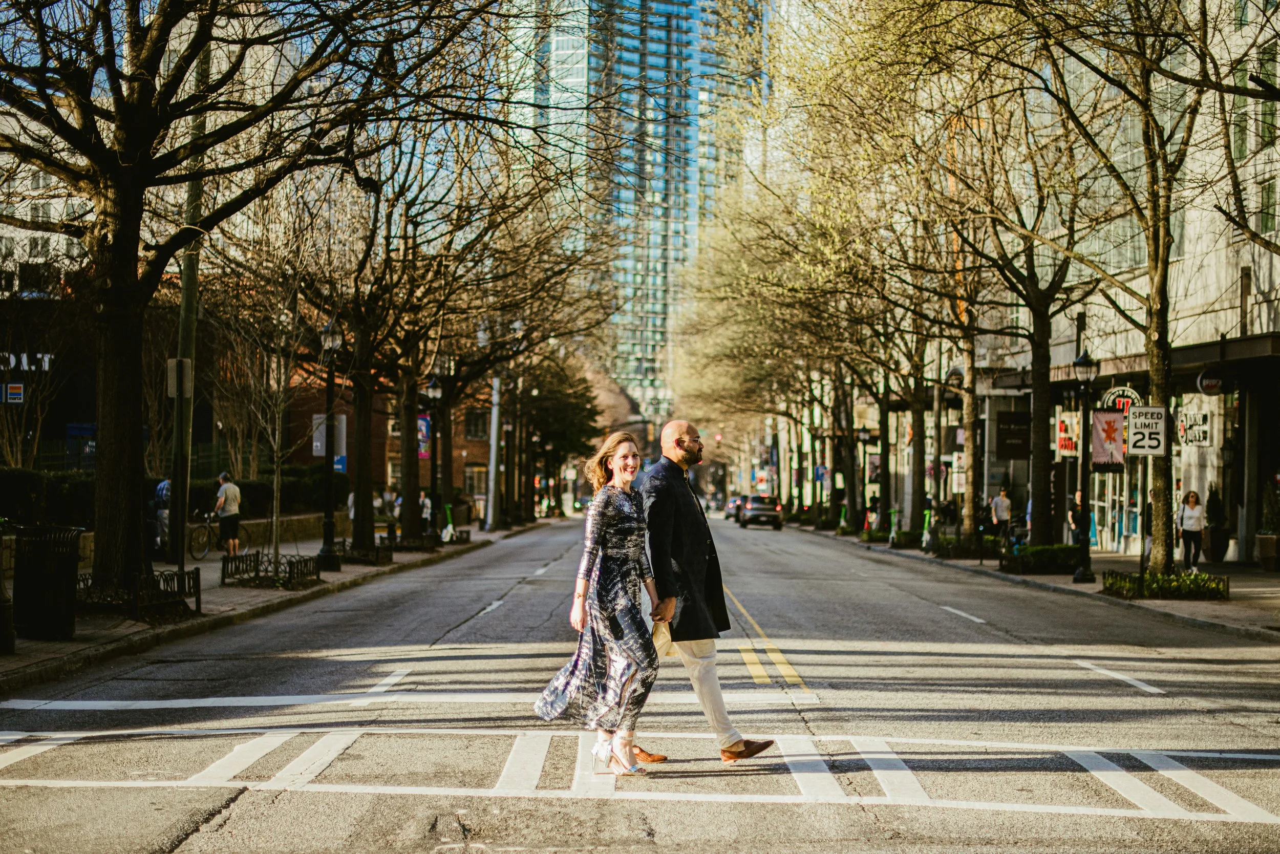 A couple crossing a city street at a pedestrian crosswalk, with leafless trees and tall buildings in the background, during daylight.