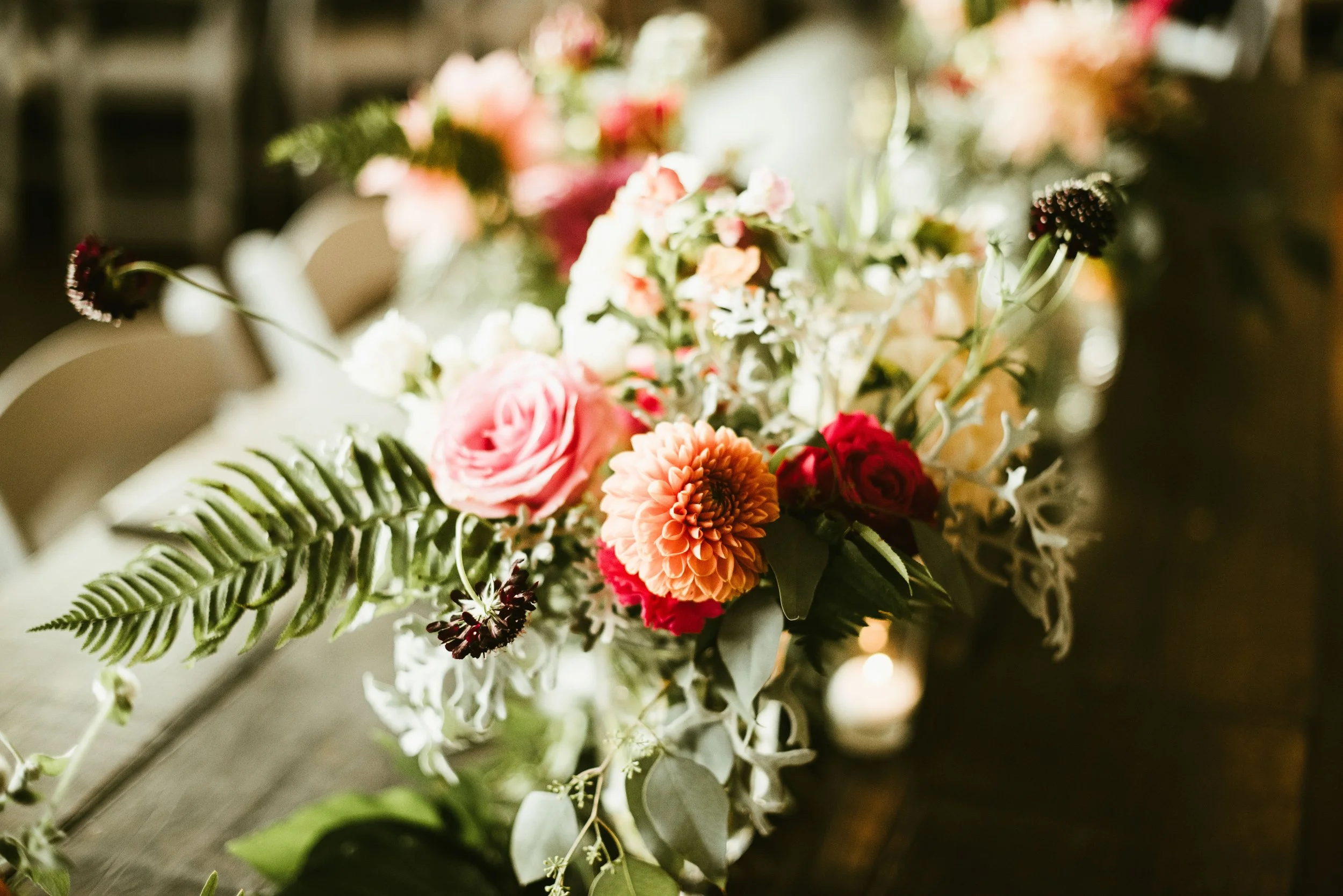 Close-up of a floral arrangement with pink, orange, and white flowers, greenery, and ferns, placed on a table in a softly lit setting.