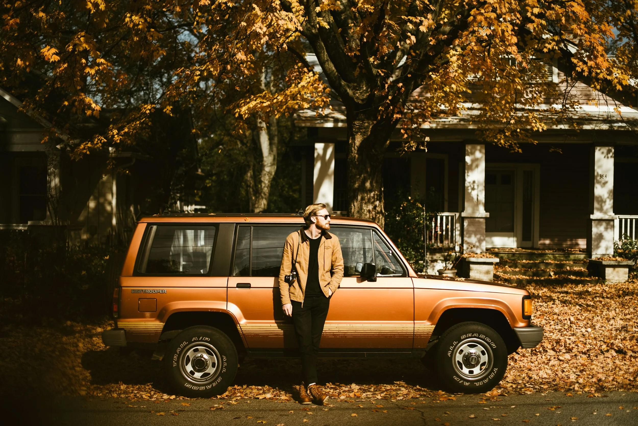 A man with a beard, sunglasses, and a tan jacket stands next to an orange SUV parked on a street. It is autumn, with yellow leaves on the trees and on the ground.