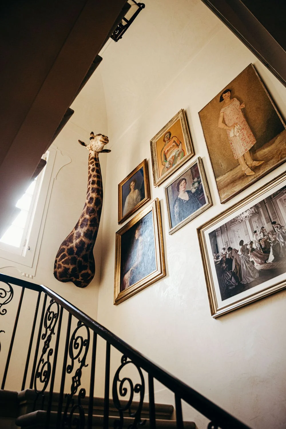A staircase with a black wrought iron railing, a wall decorated with framed vintage-style portraits, and a mounted giraffe head sculpture near a window.
