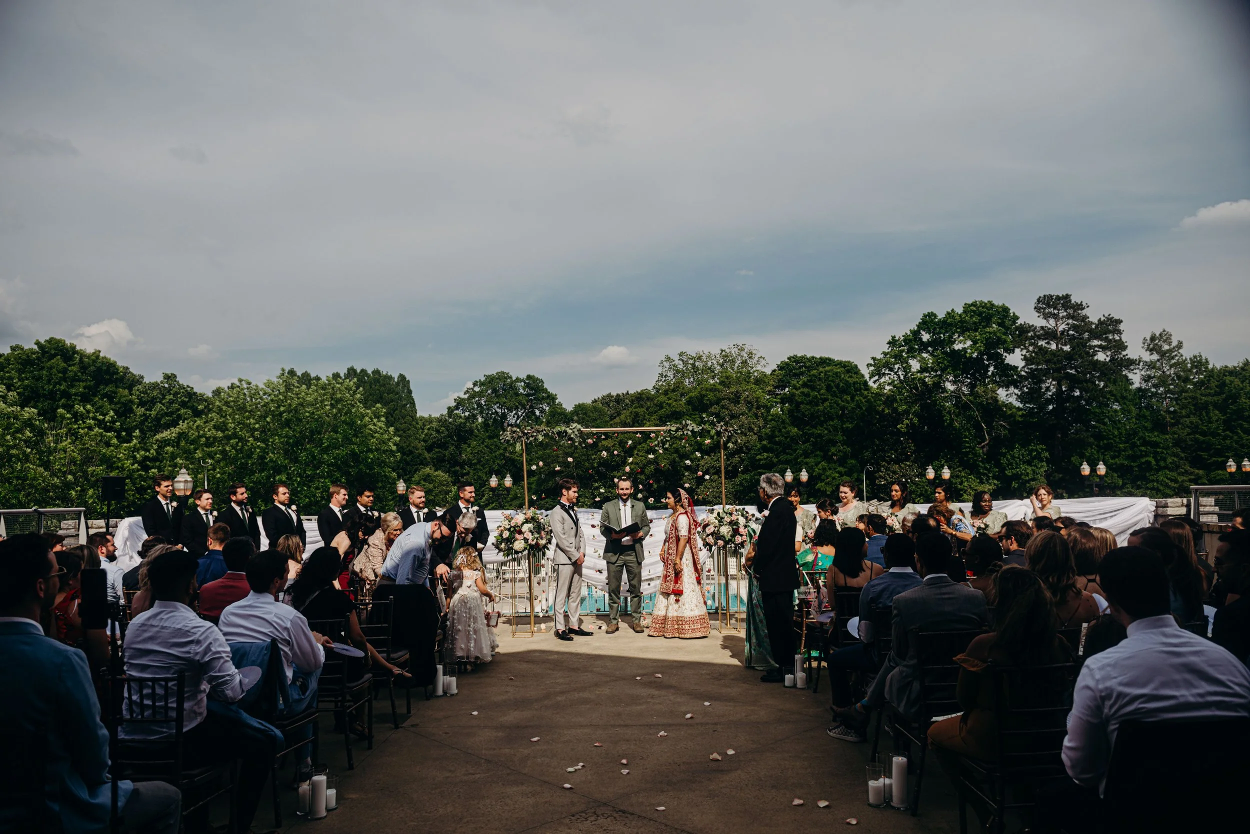 A large outdoor wedding ceremony with many guests seated, a couple standing in front of an officiant, and a wedding party dressed in formal attire, with trees and a cloudy sky in the background.