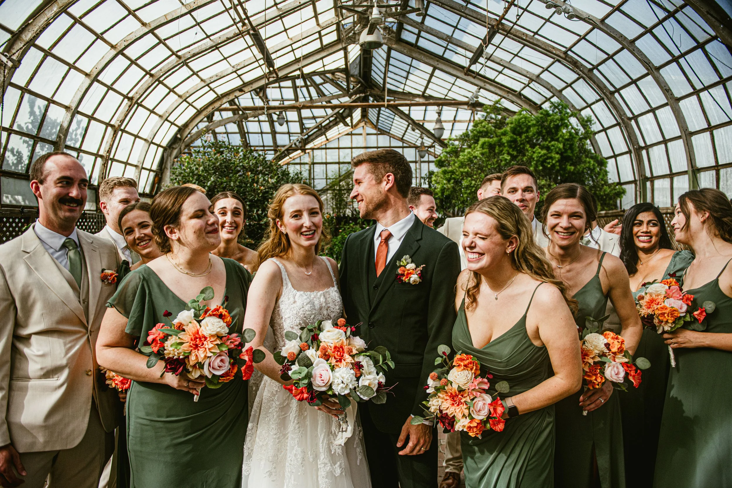 A group of people at a wedding inside a glass greenhouse, with the bride and groom in the center holding bouquets of flowers, all smiling and dressed in formal attire.