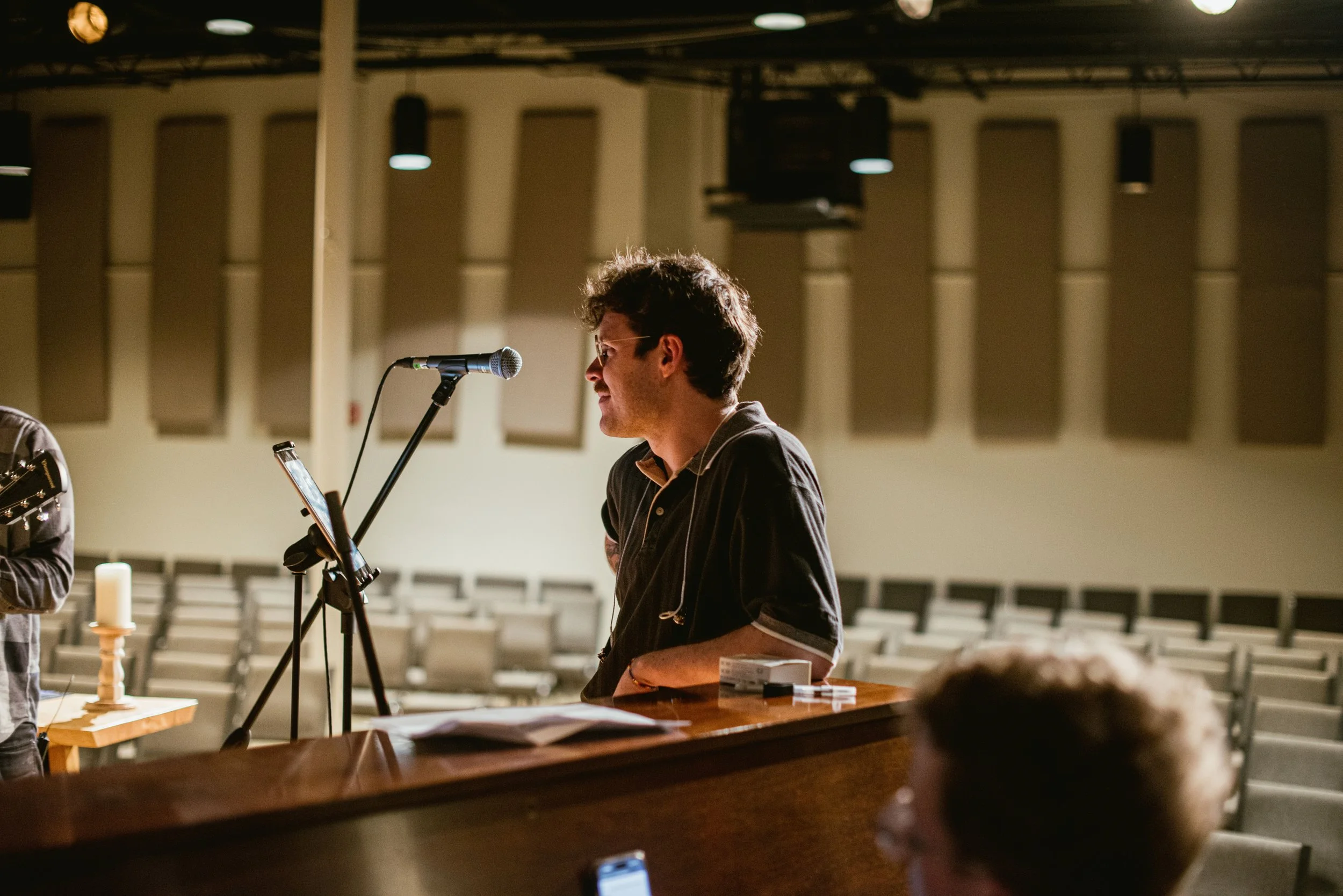A man with curly hair and glasses standing at a microphone in a large, empty auditorium, holding papers, with chairs and a candle on a wooden table nearby.