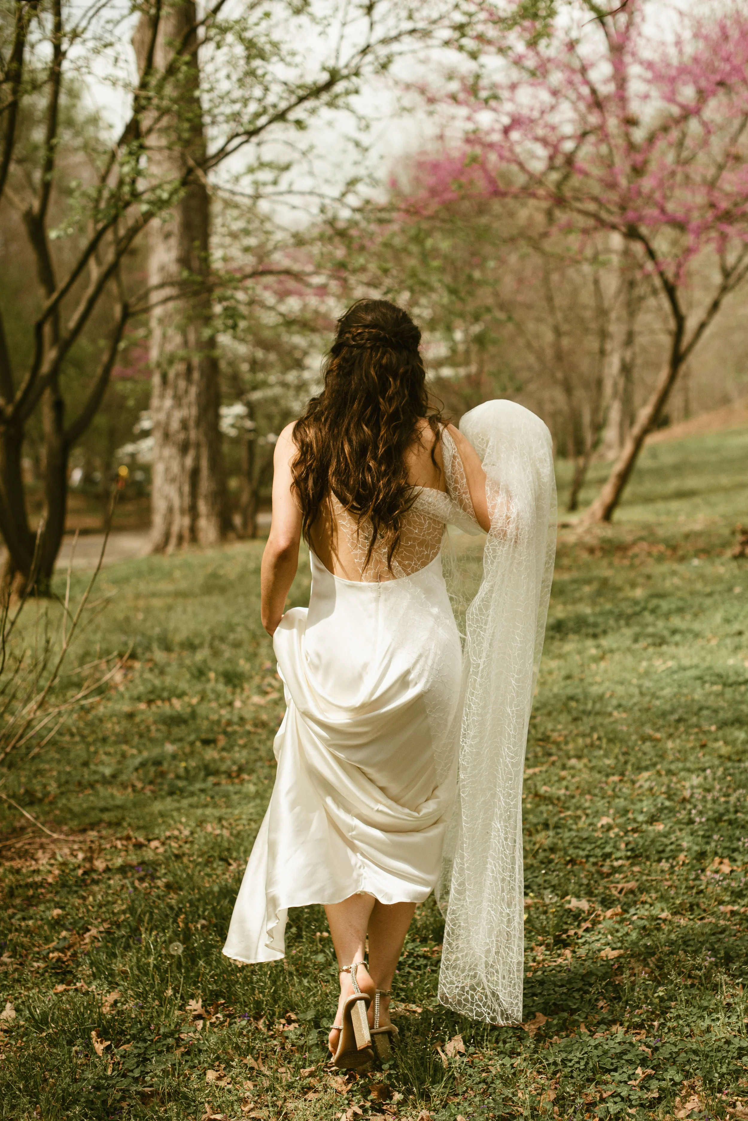 A woman with long, curly brown hair walking away in a white wedding dress through a park with blooming pink and white trees.