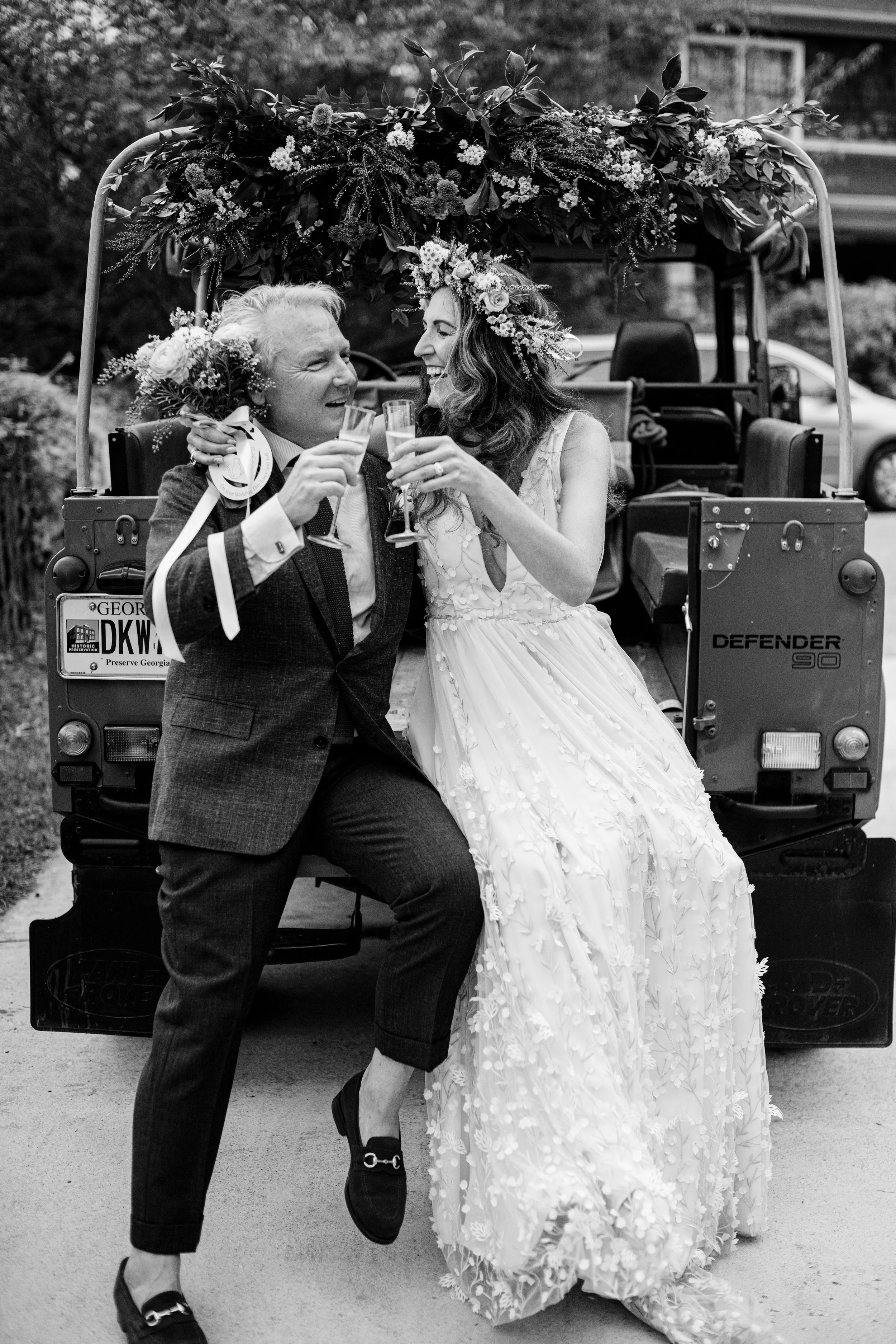A couple dancing and celebrating, with the woman wearing a wedding dress and the man in a suit, on the back of a decorated vehicle with floral arrangements.