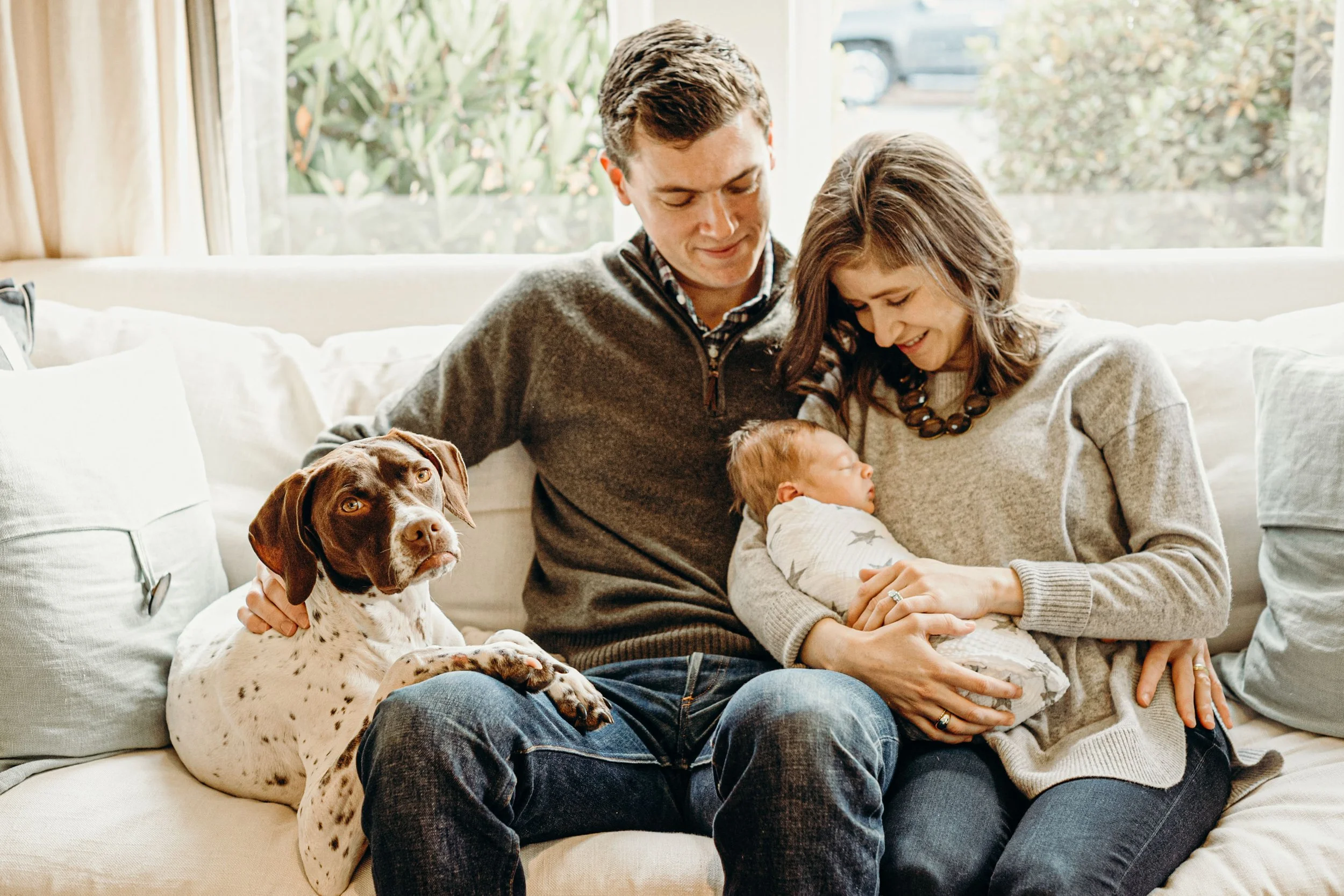 A family sitting on a couch with a newborn baby, a woman, a man, and a dog near a large window with trees outside.