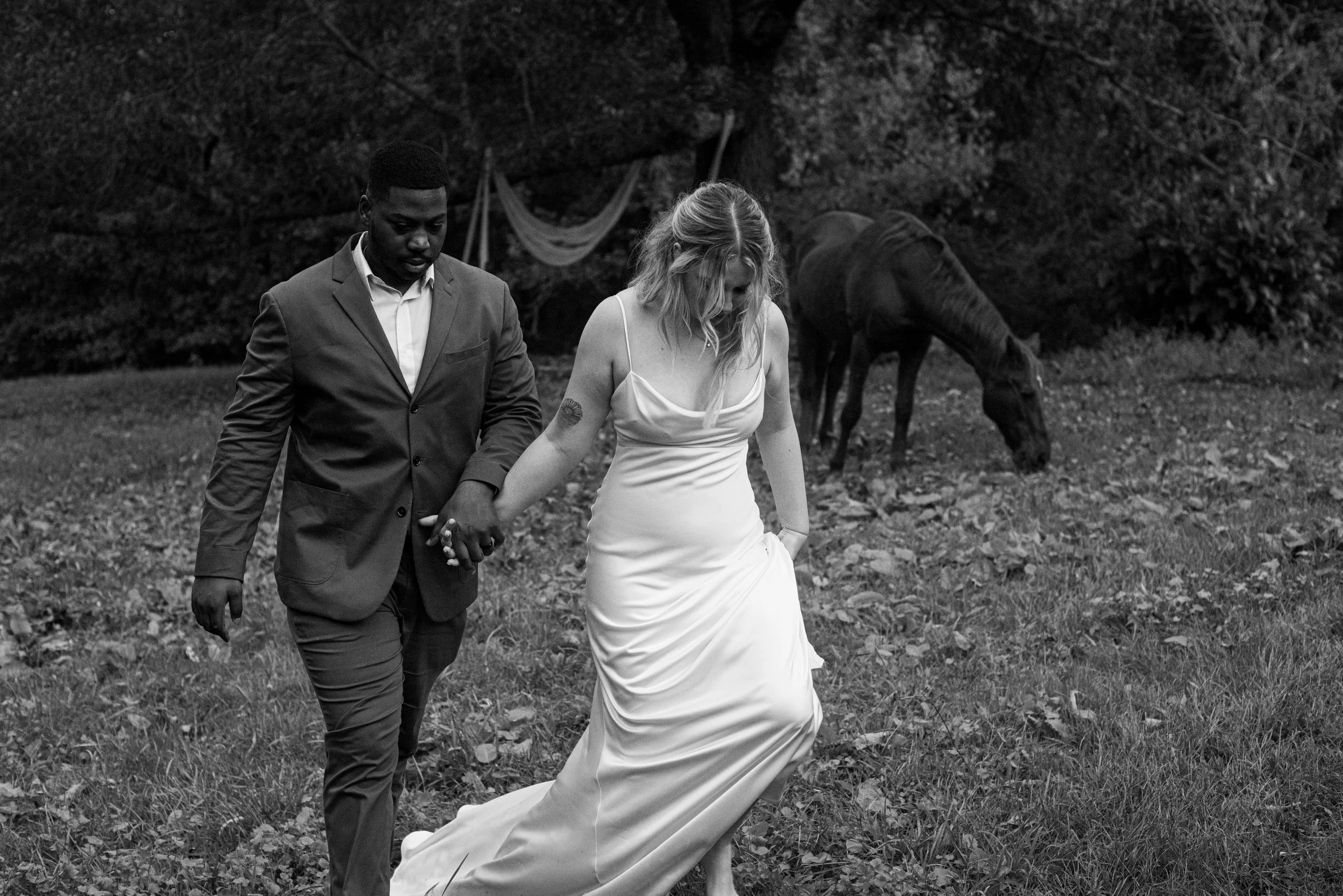 A black-and-white photo of a man and woman walking through a wooded area, holding hands. The woman is wearing a white dress, and the man is wearing a suit. There is a horse grazing in the background.