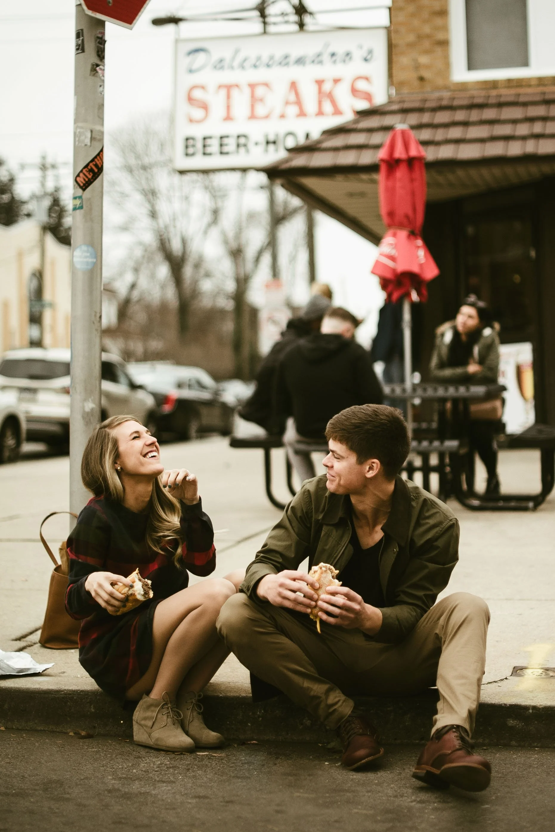 A young woman and young man are sitting on the sidewalk, smiling and eating sandwiches, with a restaurant or bar in the background. The woman has long hair and the man has short hair, both appear to be enjoying each other's company.