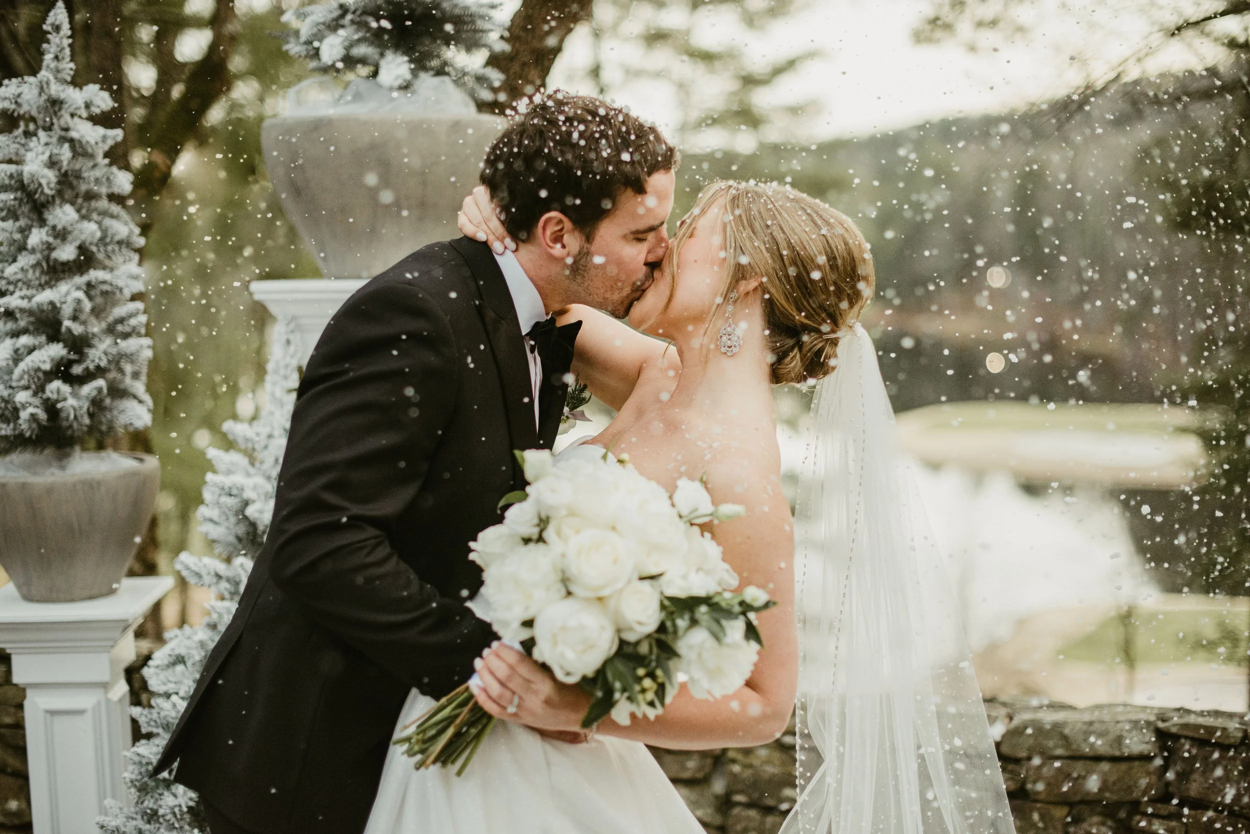 A bride and groom share a kiss during their wedding ceremony outdoors, with snow falling around them and pine trees in the background.