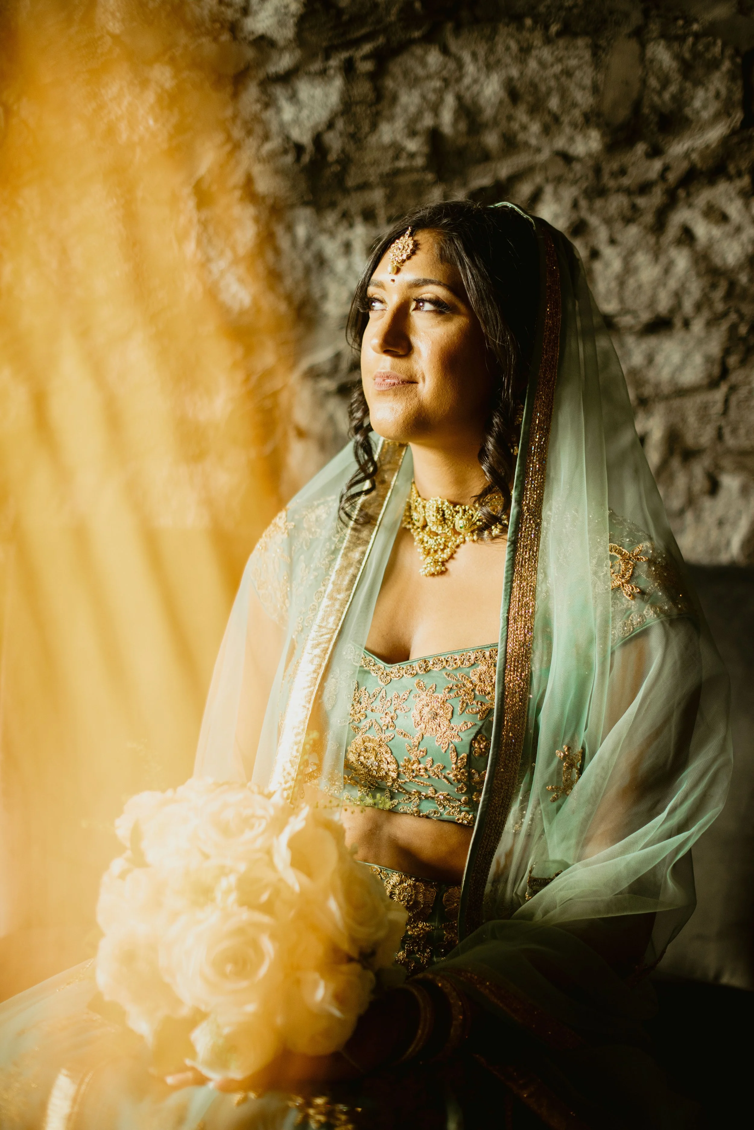 A woman dressed in traditional Indian bridal attire, wearing jewelry and a green and gold embroidered outfit, standing near a stone wall with a bouquet of white roses in the foreground.