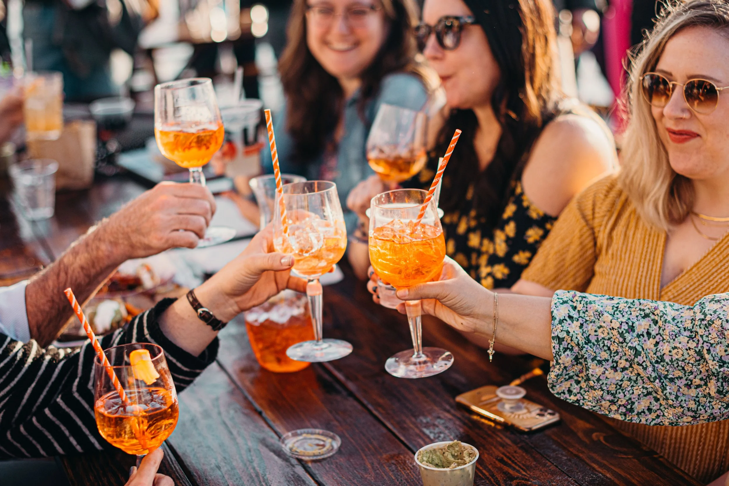 People at an outdoor gathering raising glasses of orange-colored drinks with striped straws, enjoying a social moment around a wooden table.