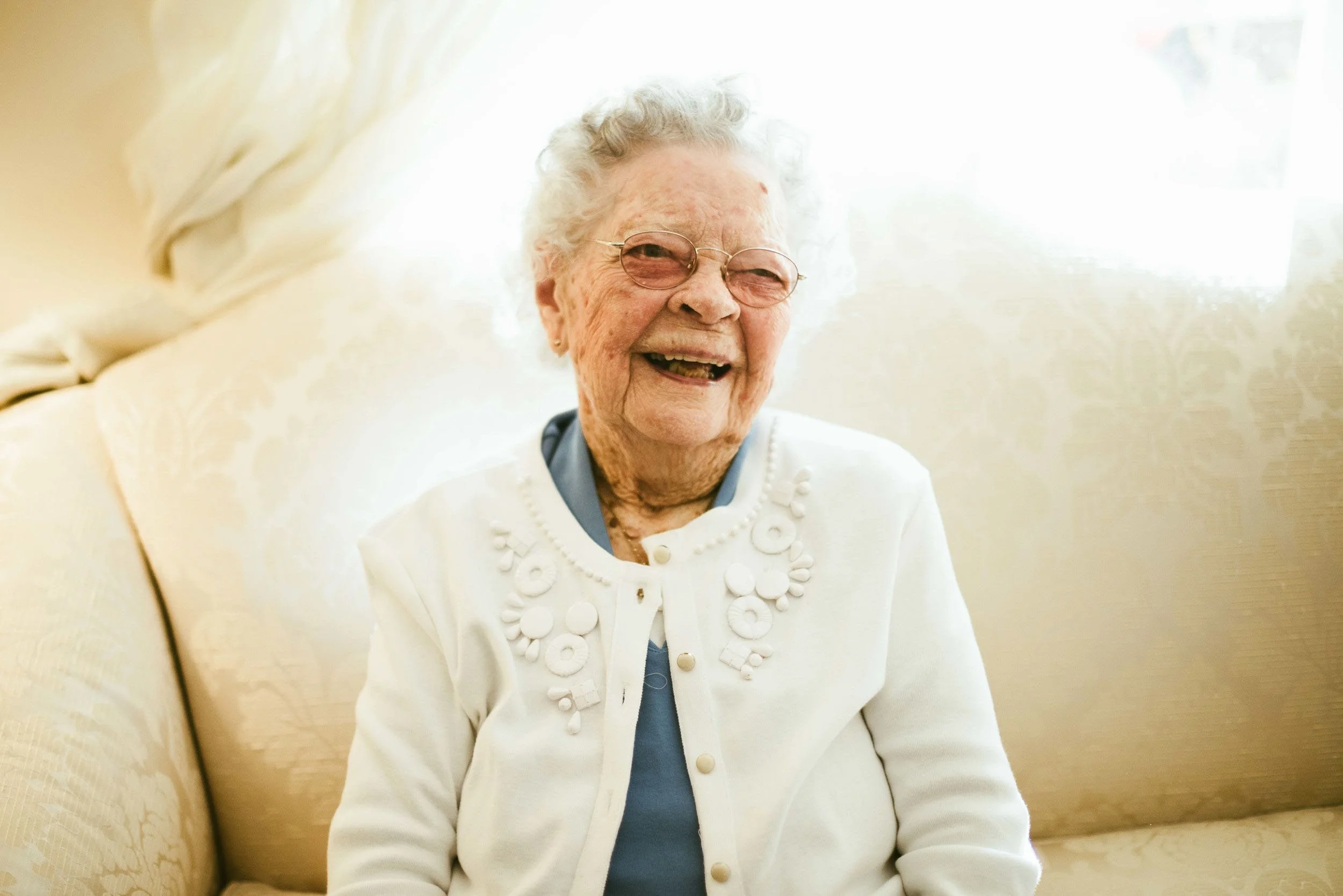 An elderly woman with curly gray hair, wearing glasses, a white cardigan with floral embroidery, and a pearl necklace, smiling and sitting on a beige couch with a light-colored curtain behind her.