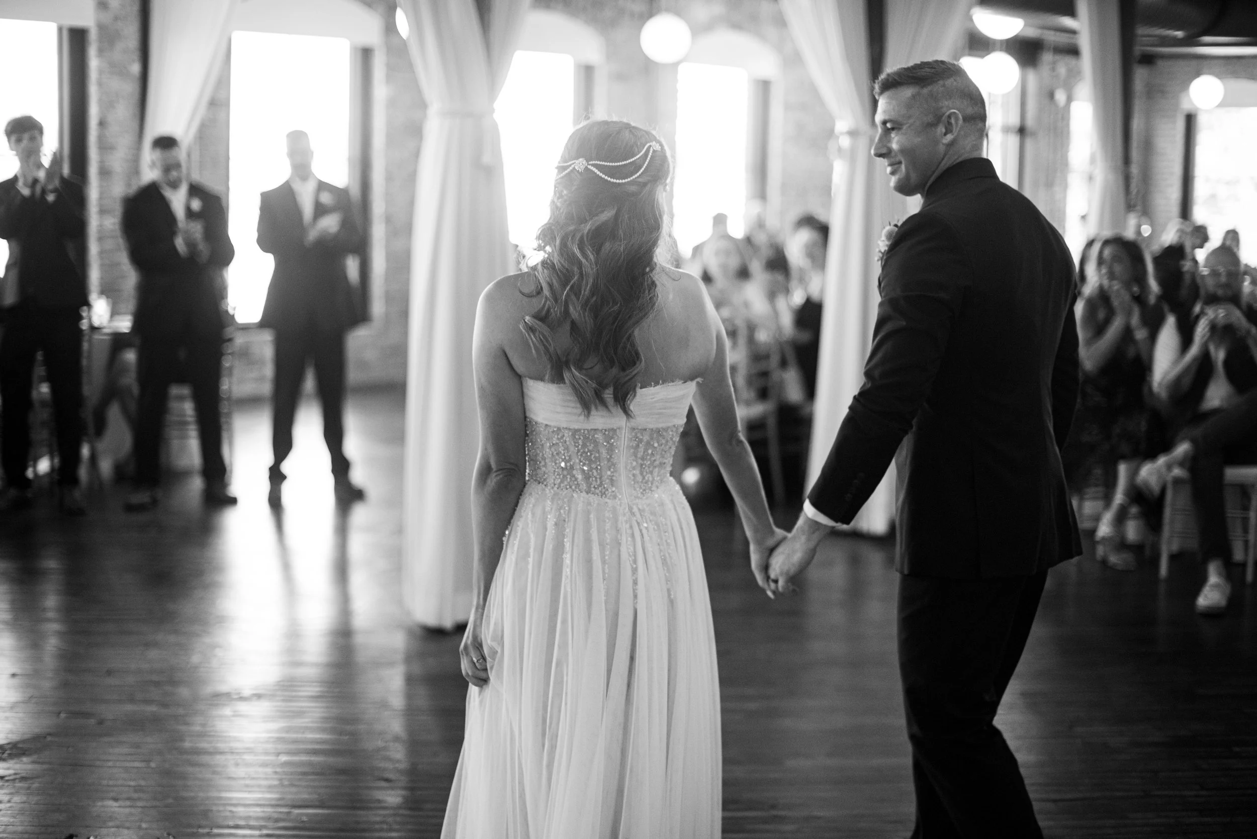 A bride and groom holding hands during their wedding dance in a decorated reception hall with guests watching.