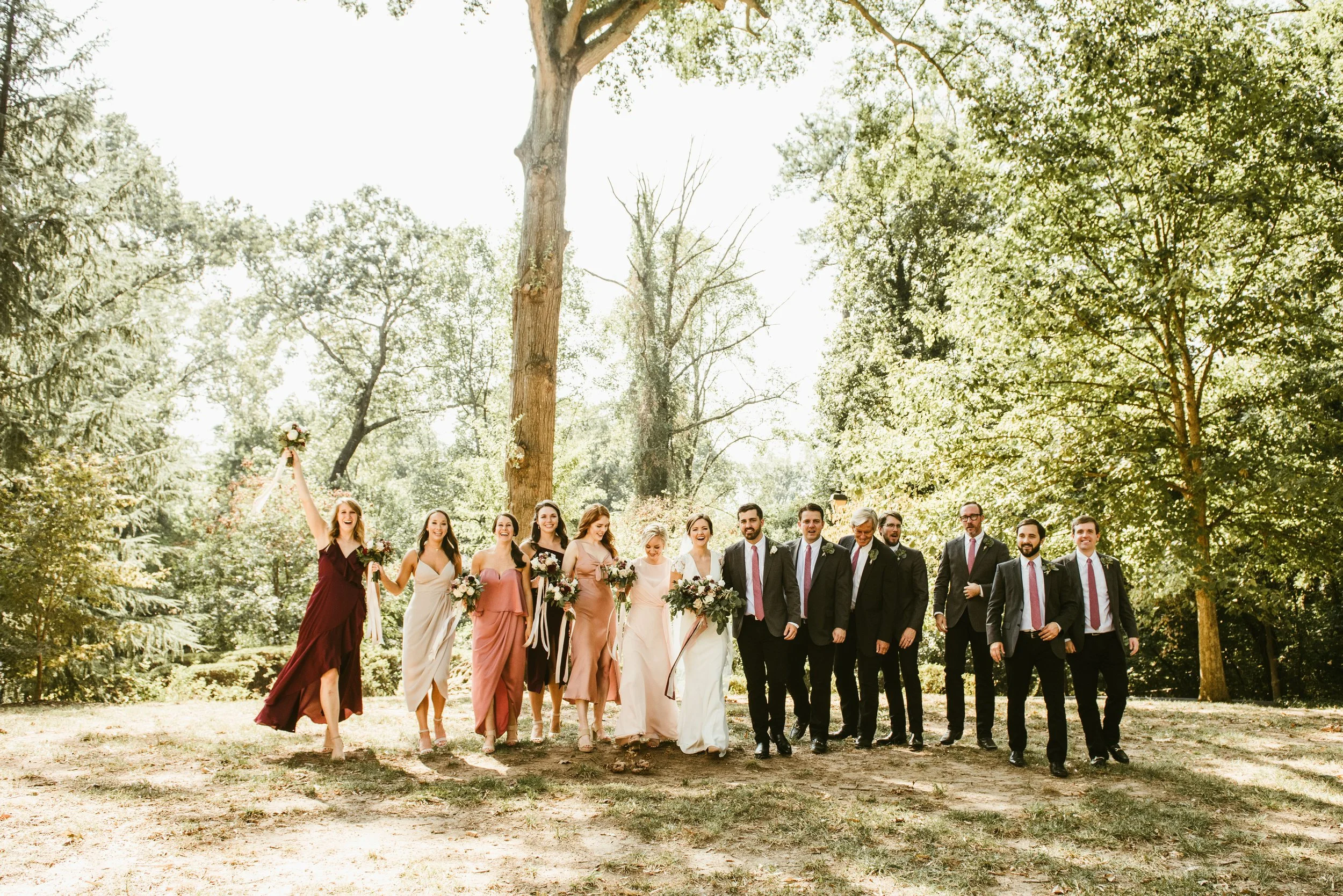 A group of wedding guests walking outdoors on a sunny day in a park, surrounded by tall trees and green foliage.