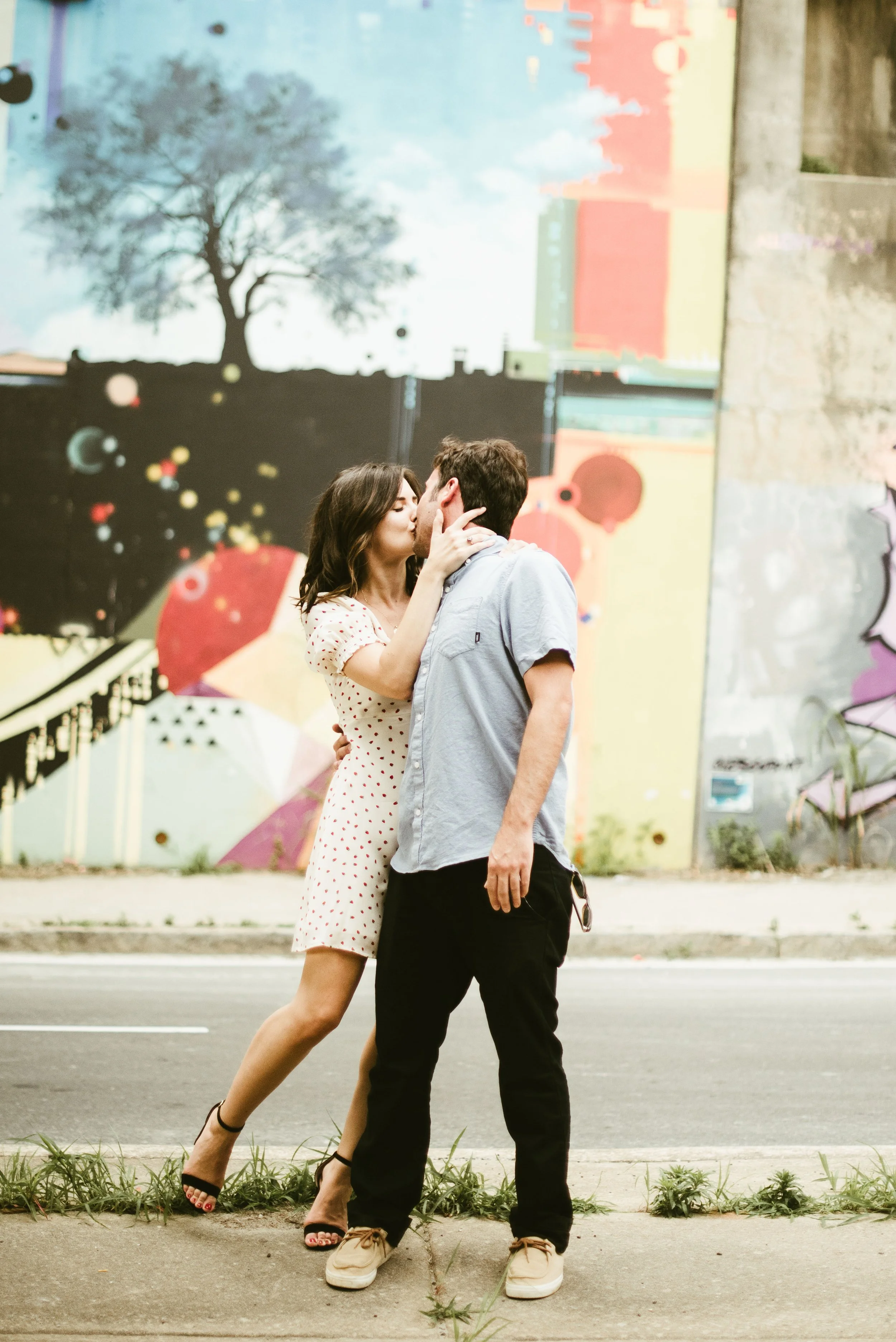 A couple sharing a kiss while standing on a street in front of a colorful graffiti wall.