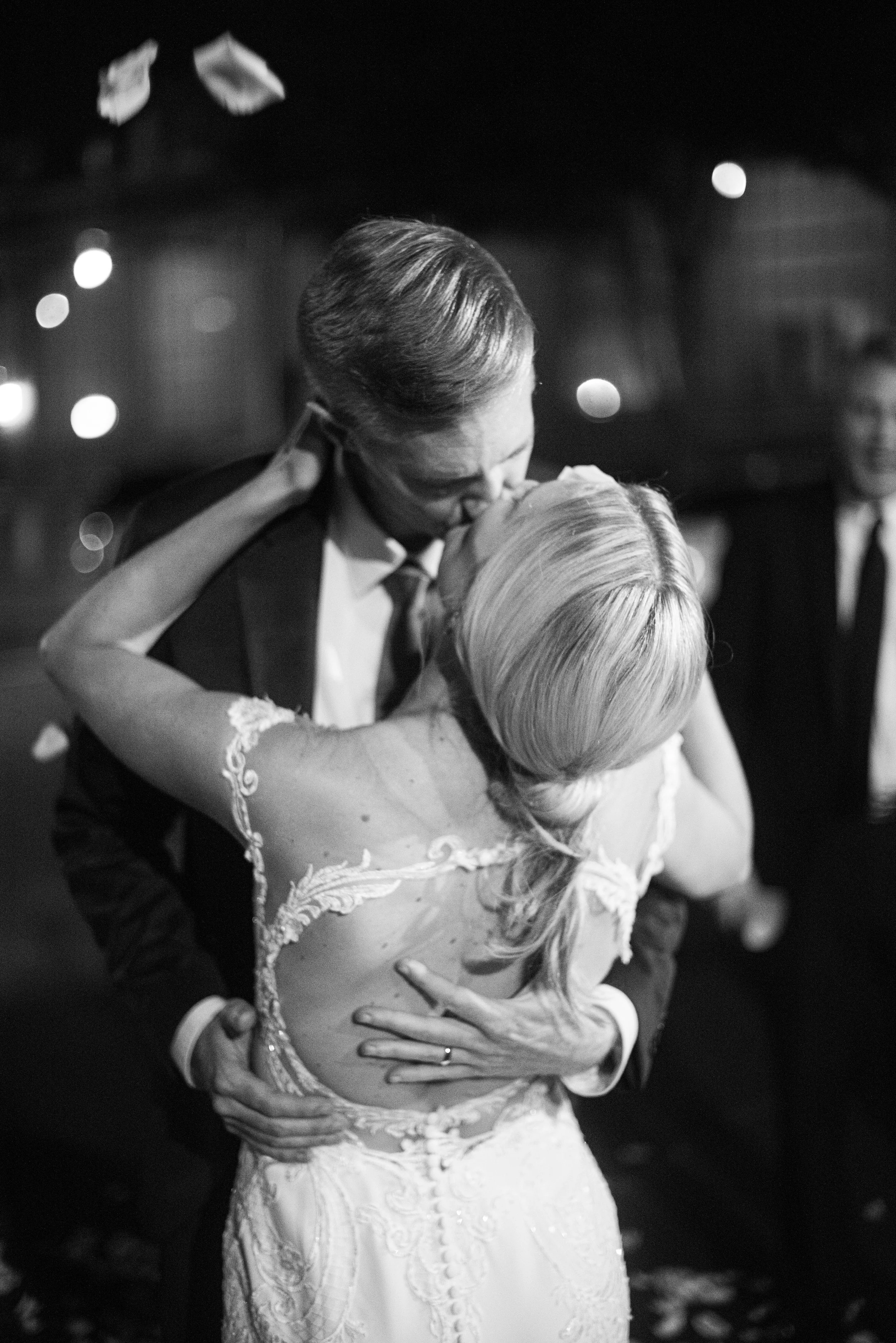 A black and white photograph of a bride and groom sharing a dance. The bride has blonde hair styled in loose waves and is wearing a lace wedding dress. The groom has slicked-back hair and is dressed in a suit with a tie. They are embracing closely, w