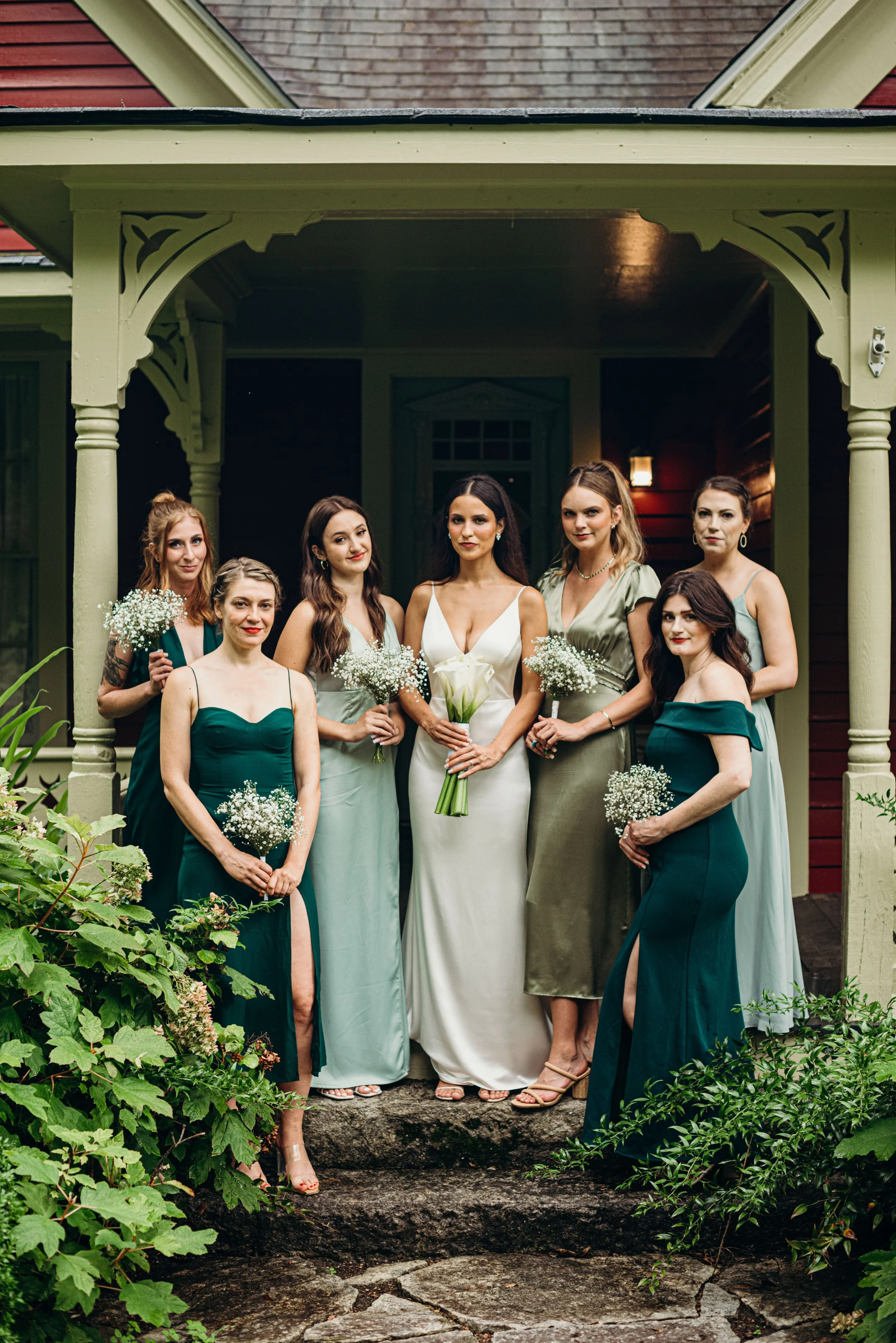 Group of seven women in formal dresses standing on stone steps outdoors, with some holding bouquets of white flowers, in front of a house with a porch and red and brown siding, during evening or dusk.