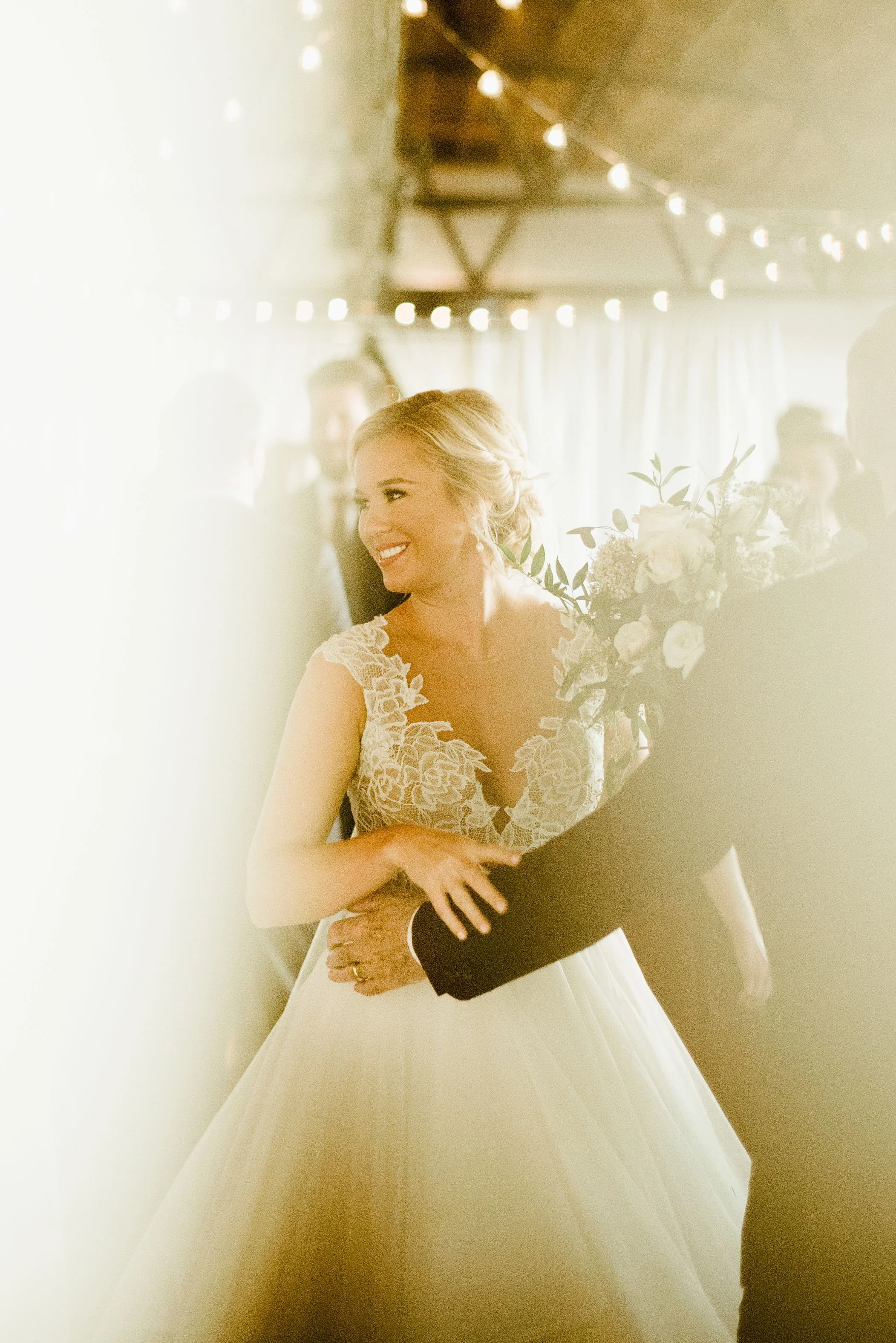 A bride with blonde hair in an updo, wearing a lace wedding gown, cheerfully embracing a person in a black tuxedo at her wedding reception.