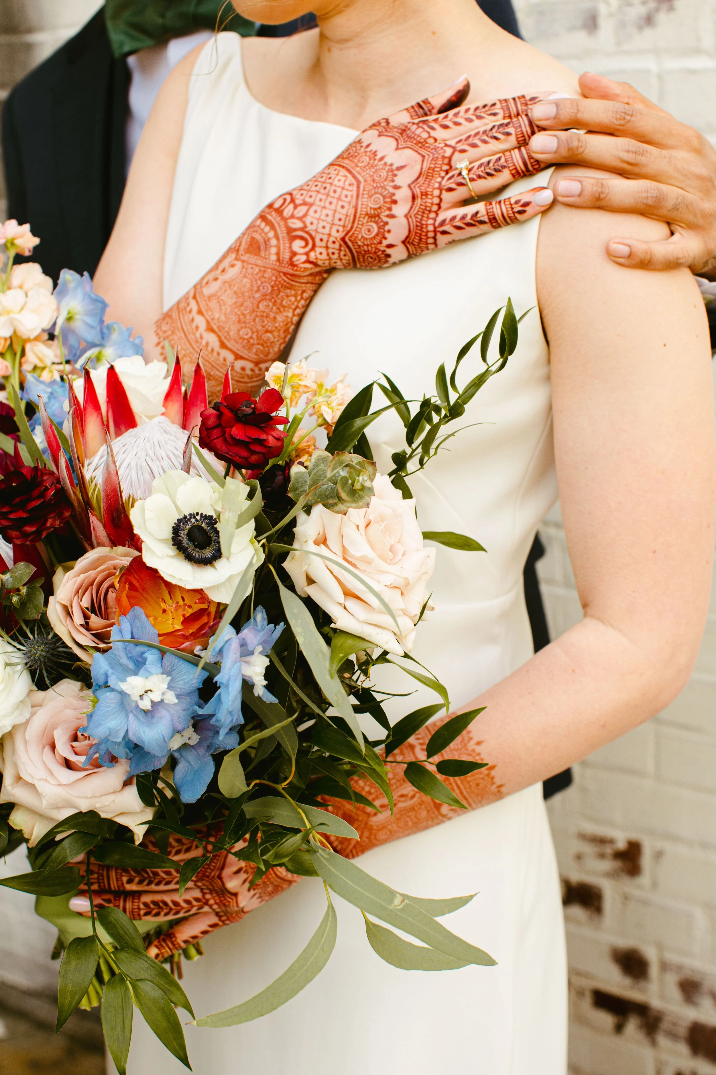 A bride is holding a large bouquet of various colorful flowers, and a groom is embracing her from behind with his hands on her shoulder. The bride is wearing a white dress, and the groom is in a black suit with a white shirt. The bride has henna on h