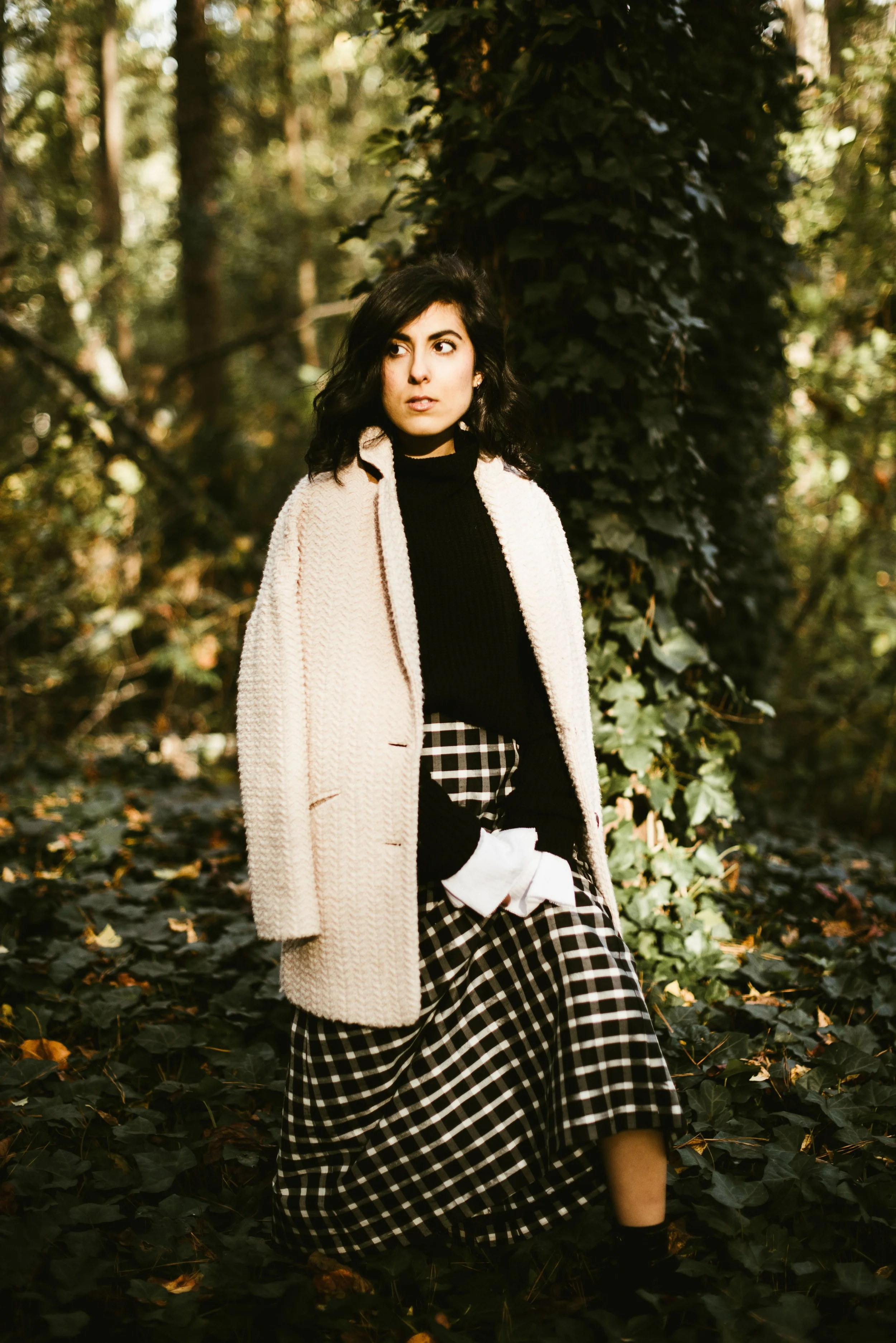 A woman with dark hair and a beige coat standing in a forest with green ivy and foliage, looking to the side.