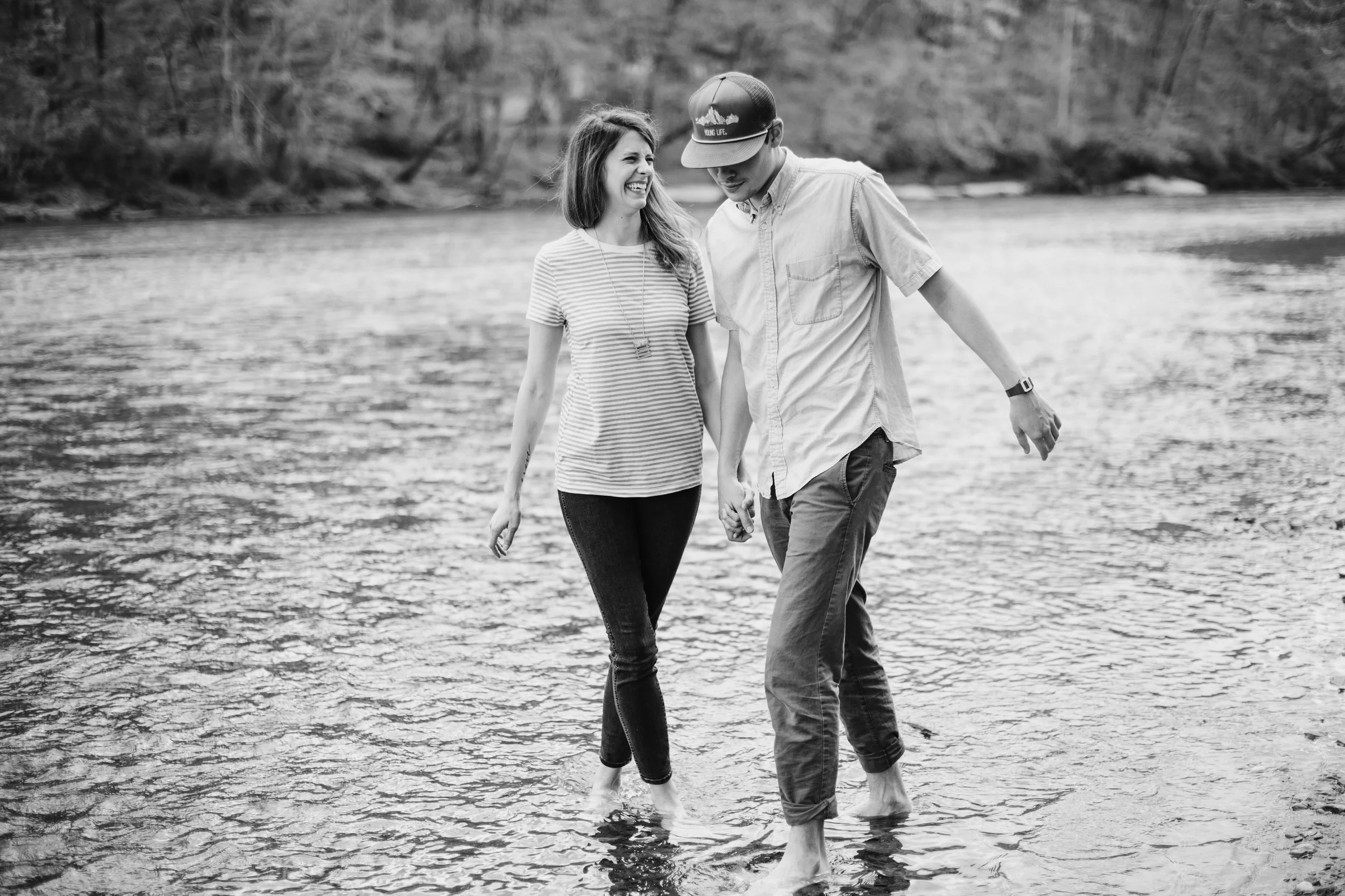 A black-and-white photo of a young man and woman walking barefoot in shallow water near a river, holding hands and smiling, with trees in the background.