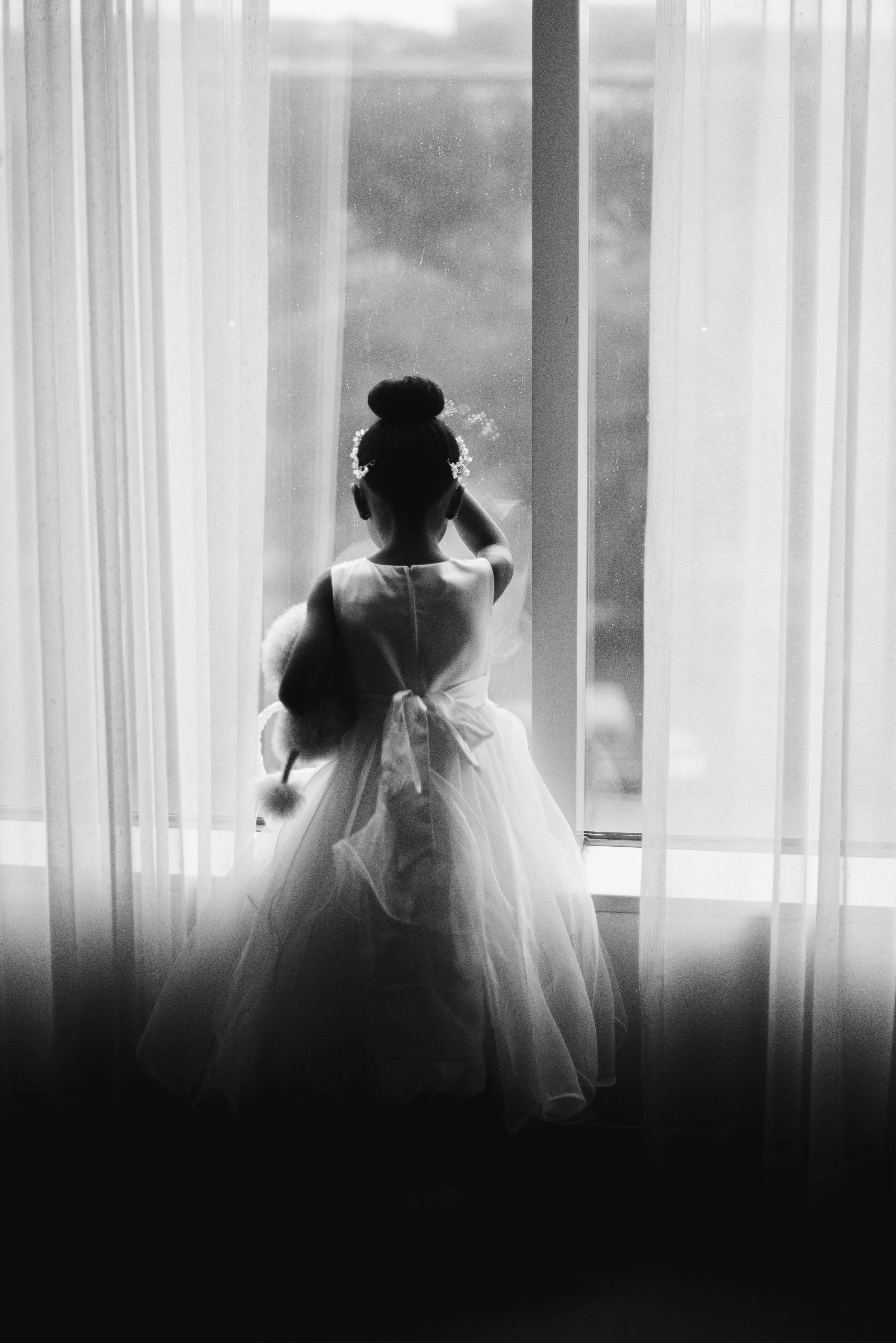 A young girl in a wedding dress standing in front of a window with sheer curtains, looking outside.