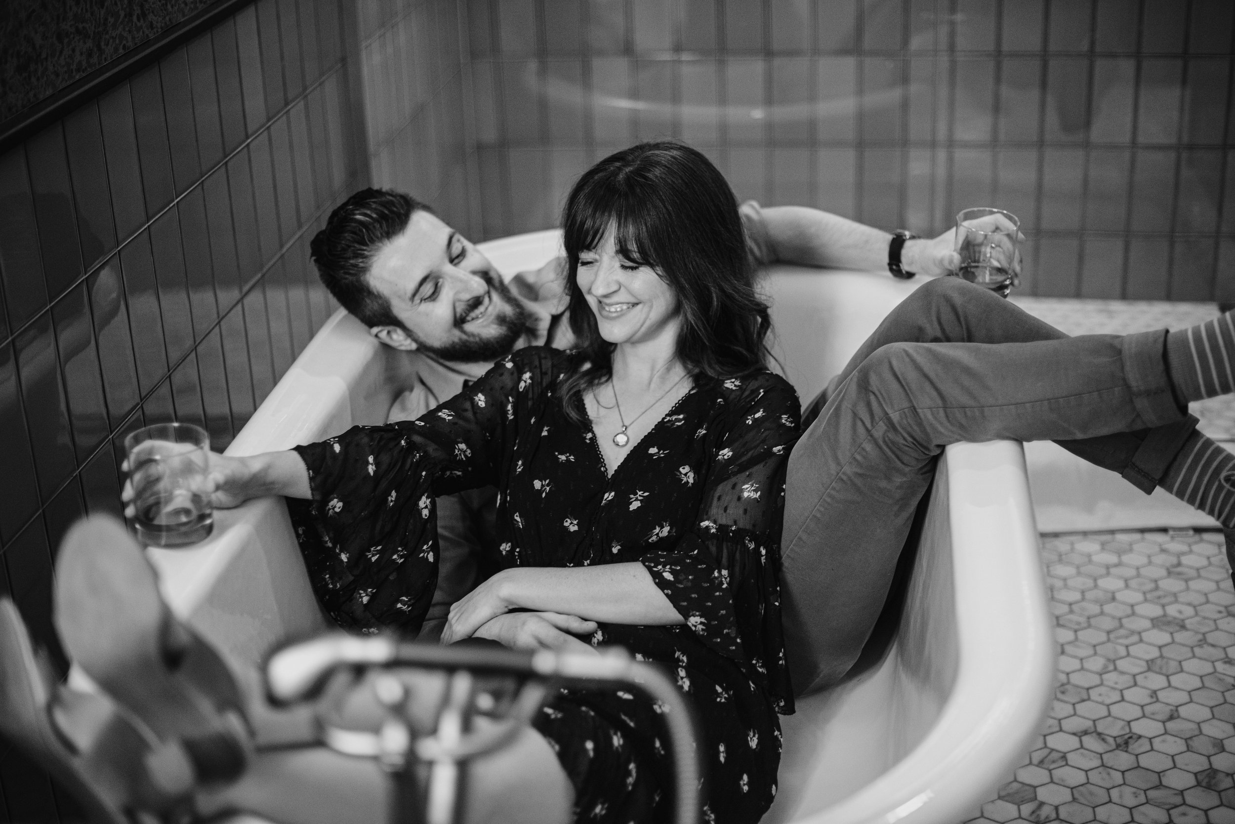 A couple smiling and enjoying a drink together in a bathtub in a bathroom.