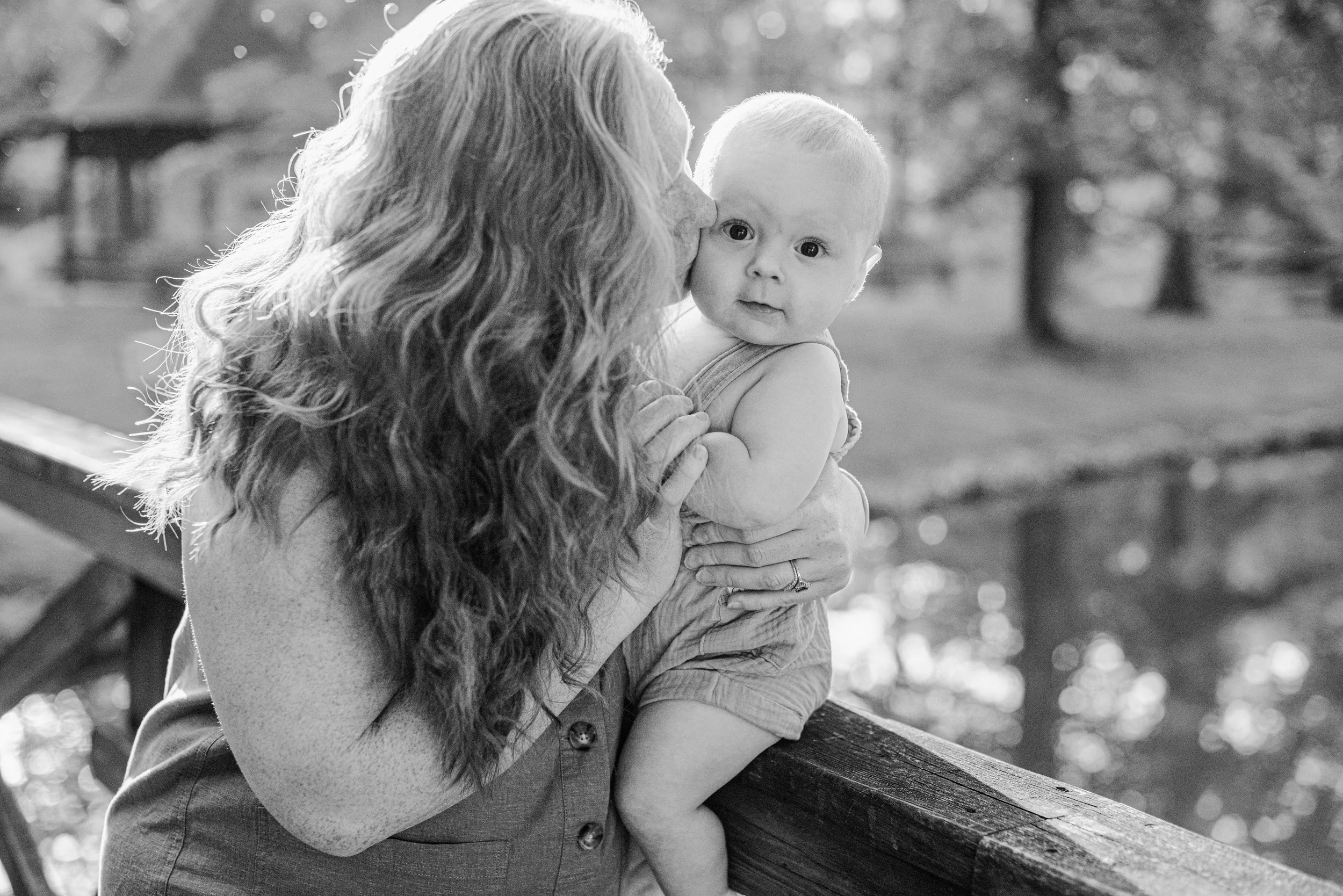 A mother with long curly hair holding a young child on a park bench, with trees in the background, in black and white.