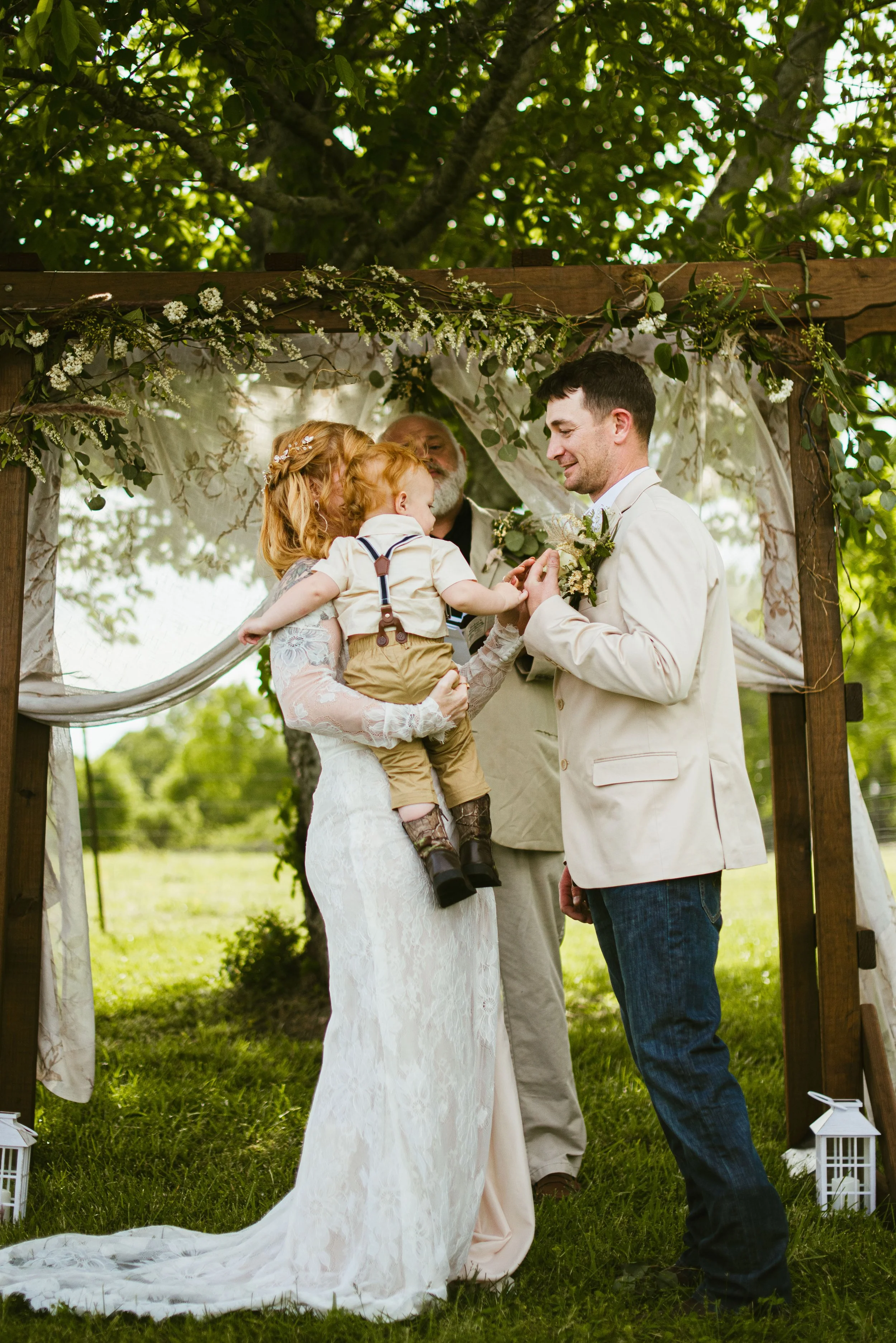 A wedding ceremony outdoors with a bride holding a young boy, a groom, and an officiant under a decorated wooden arch.
