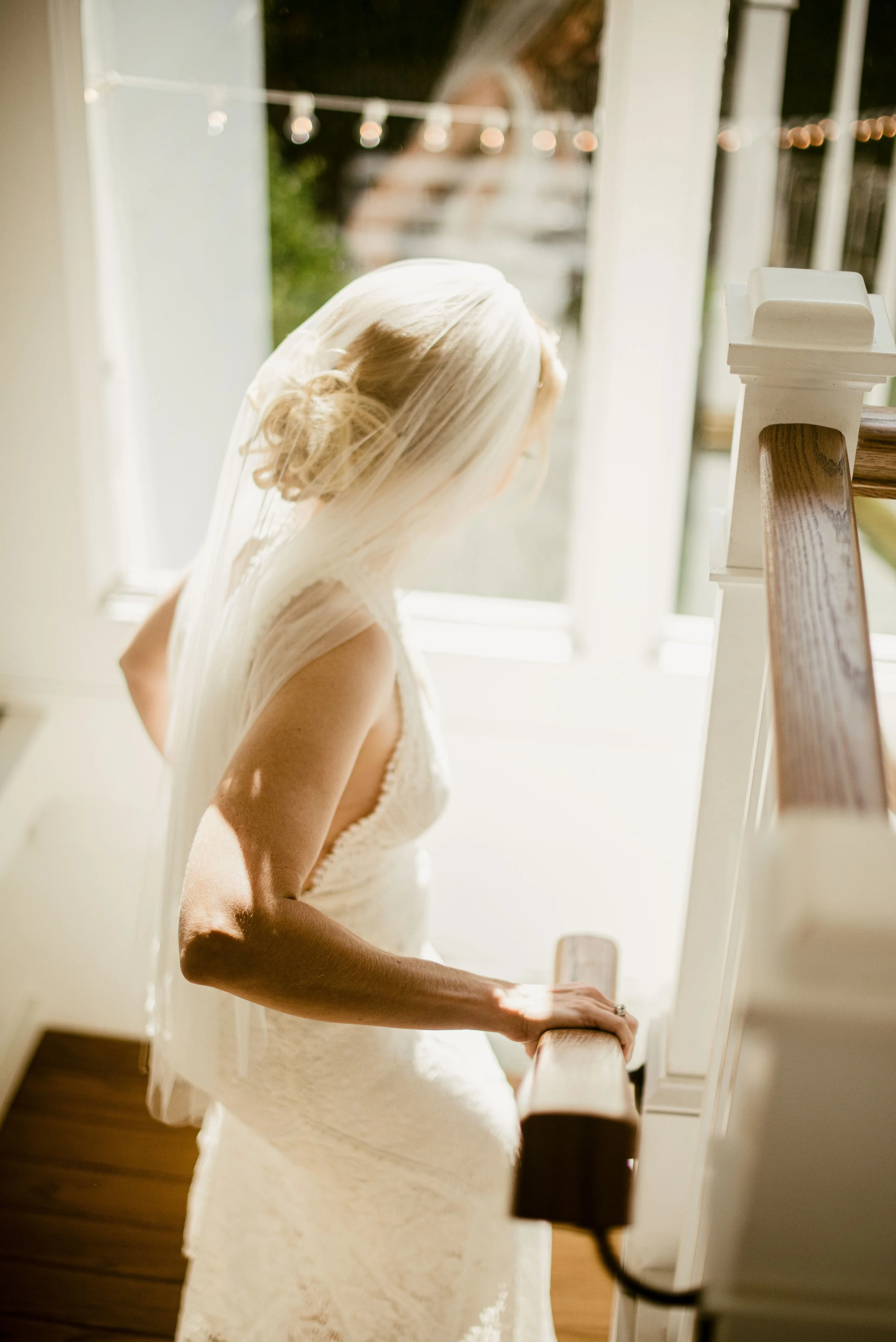 A woman in a white lace dress with blonde hair styled in an updo, standing by a staircase with her hand resting on the railing, looking out a window with blurred outdoor string lights visible.