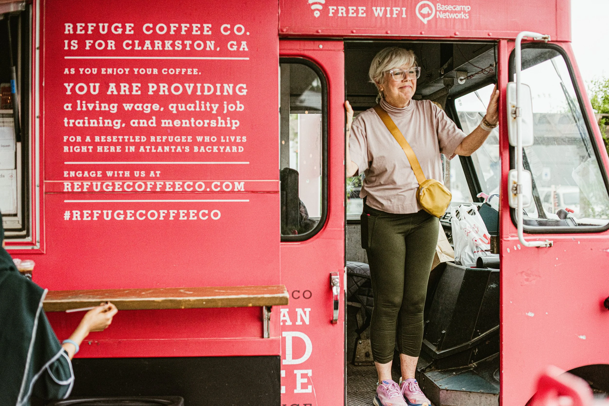 A woman standing inside a red food truck with a sign promoting refugee support and free Wi-Fi, wearing glasses, a beige shirt, black pants, pink shoes, and carrying a yellow bag.