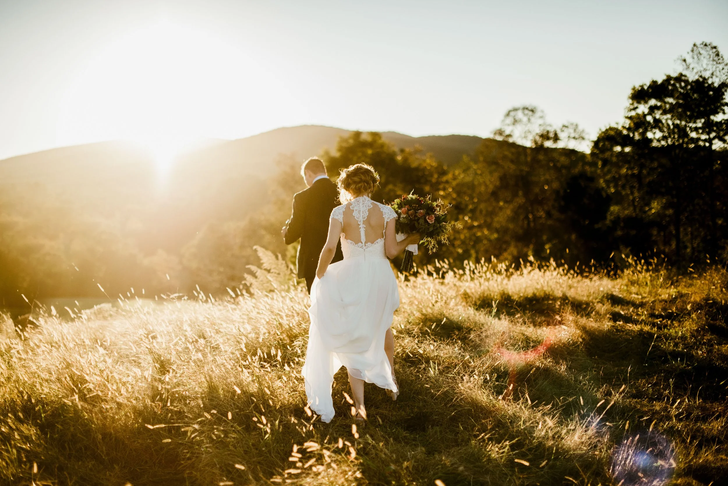 A bride and groom walking through a grassy field at sunset, with the bride holding a large bouquet of flowers.