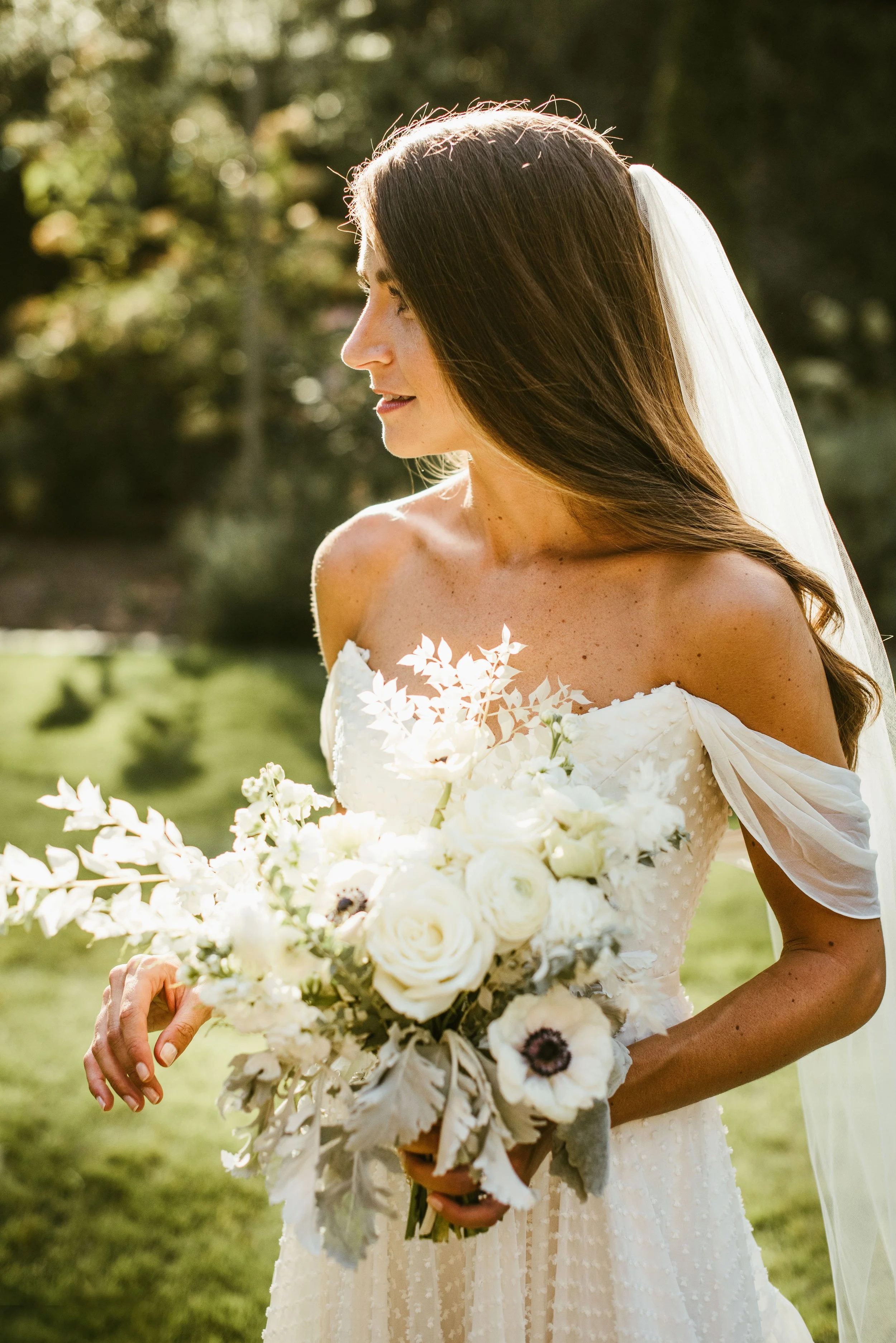 A bride with long brown hair wearing a white wedding dress holding a large bouquet of white flowers outdoors during daytime.